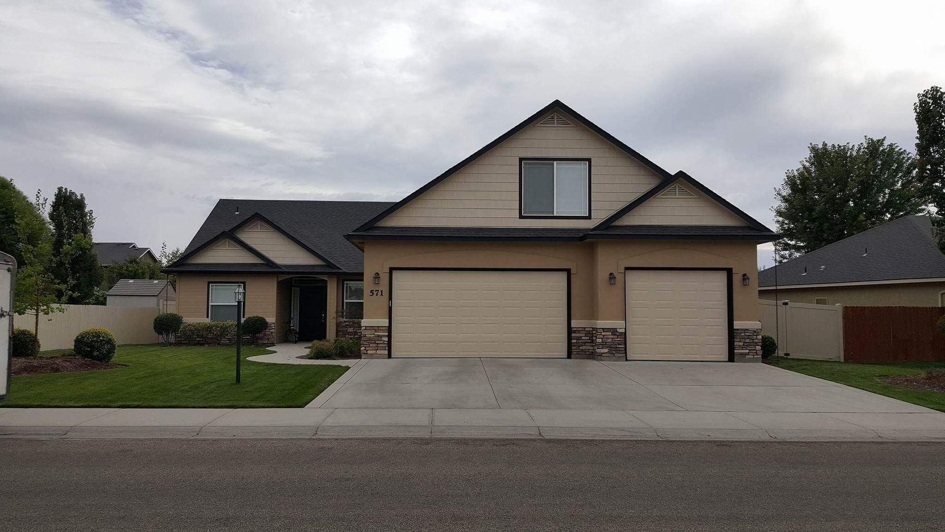 Tan house with two-car garage and dark gray roof on a cloudy day. Front yard with green grass.