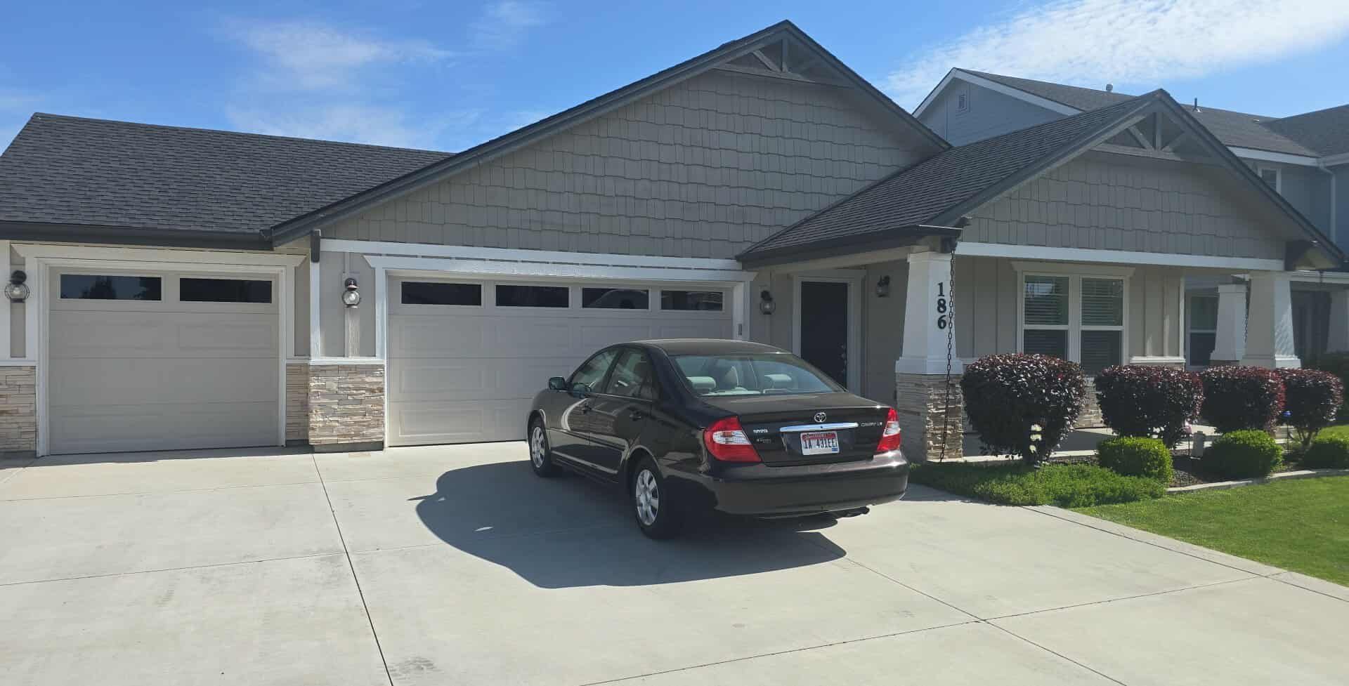A black car parked in front of a gray house with a two-car garage. The house has a dark roof and a green lawn.