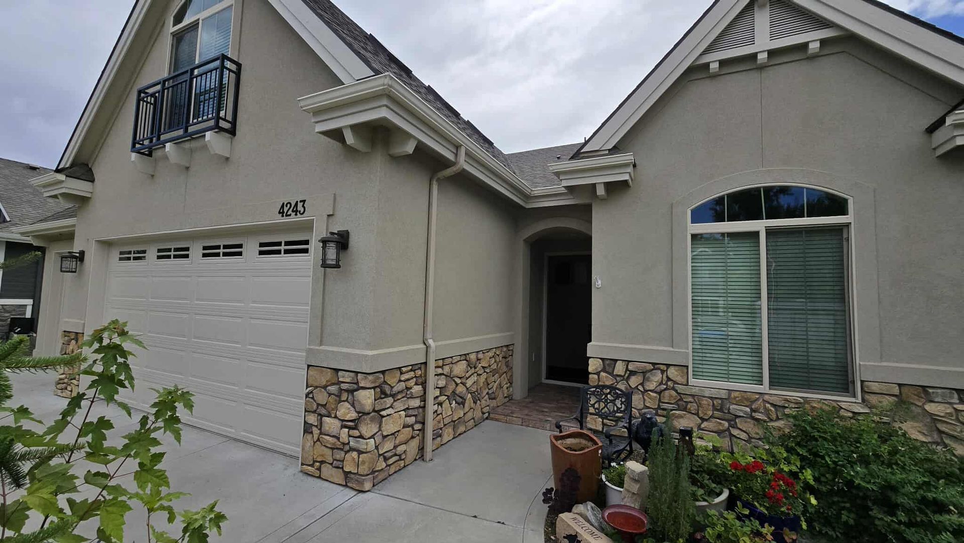 Exterior view of a tan stucco house with stone accents, a white garage door, and arched windows. The front door is open, and landscaping is visible.