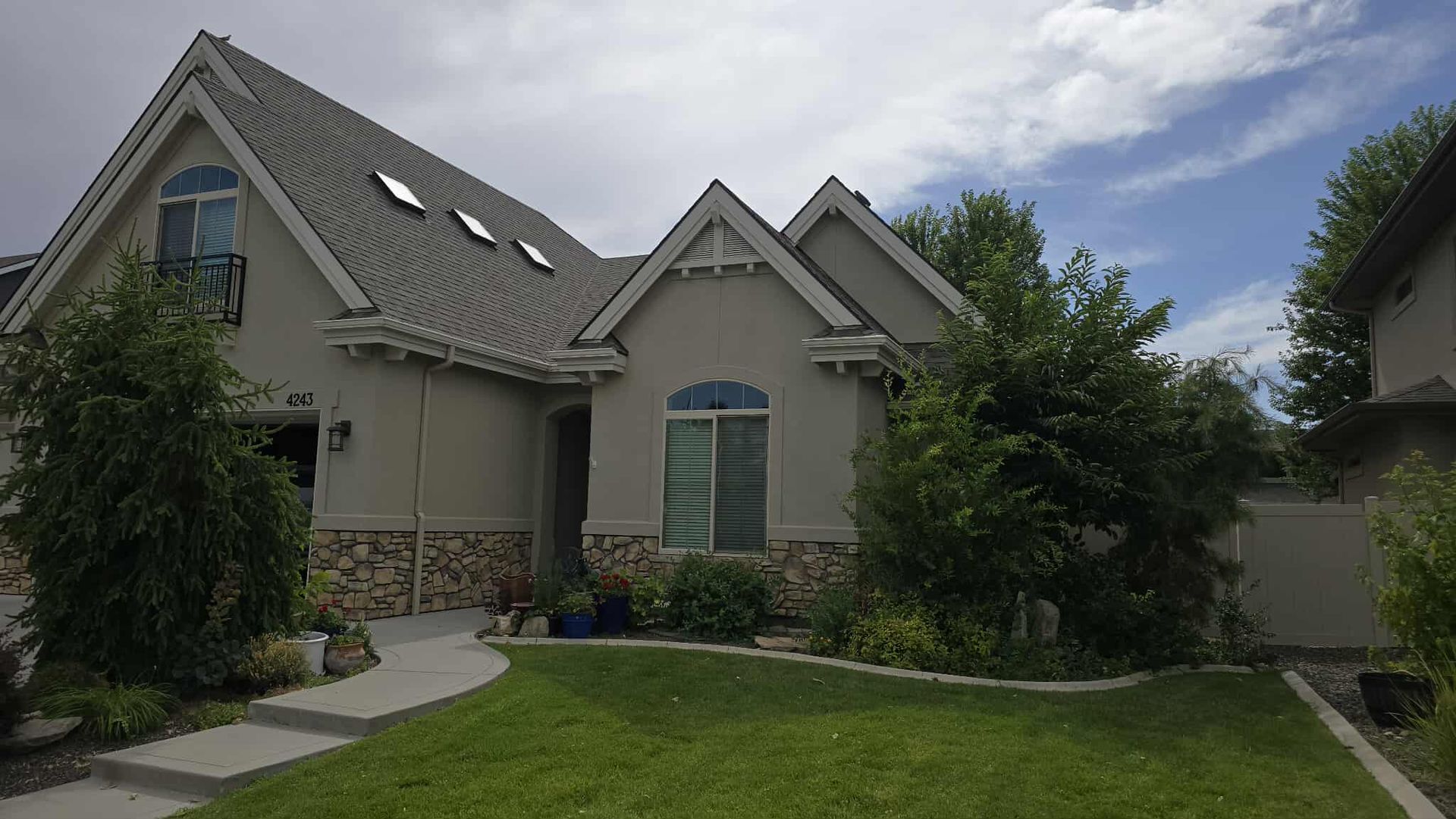 Tan stucco house with gray roof, green lawn, and cloudy sky. The front of the house has a walkway leading to the front door.