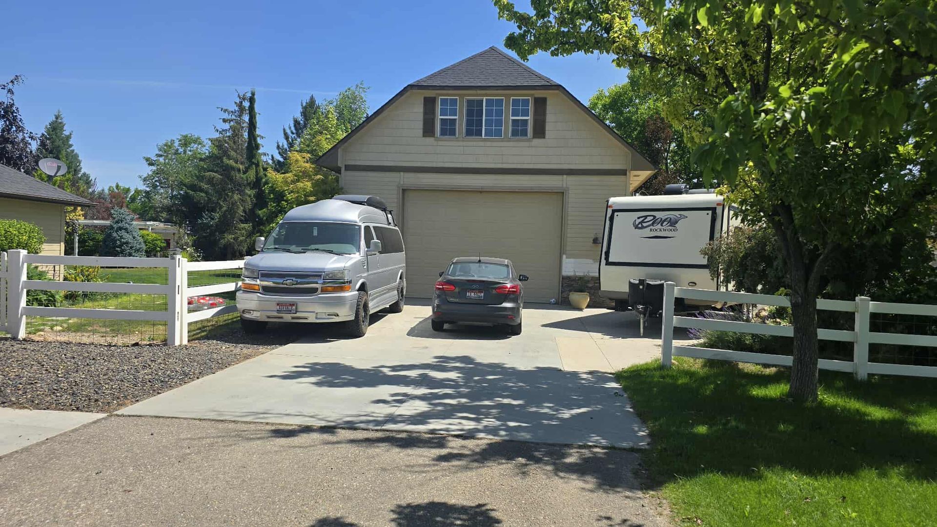 A driveway with a silver van, black car, and RV parked in front of a two-car garage with a dormer on a sunny day.