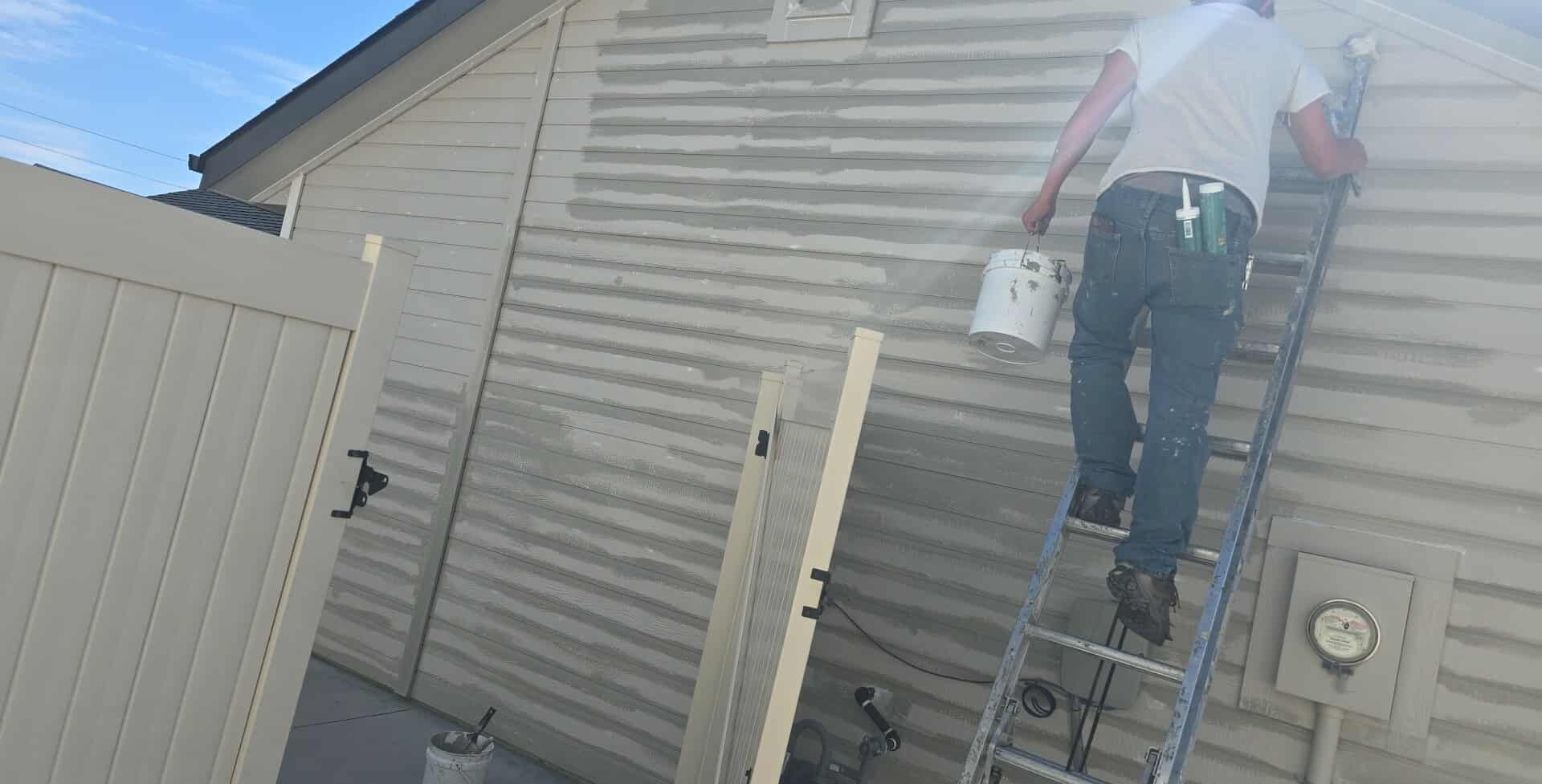 A person climbs a ladder to paint siding on a house. They hold a bucket of paint, with a neutral-toned fence nearby.
