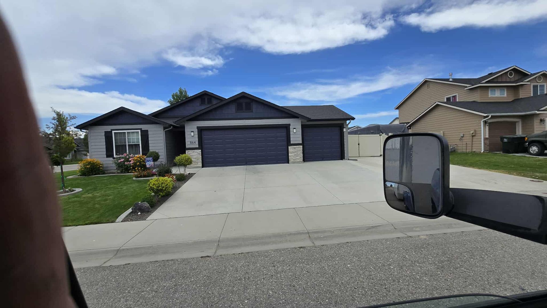 A single-story house with a dark gray garage door and trim, with a lawn and blue sky in the background. The photo is taken from inside a vehicle.