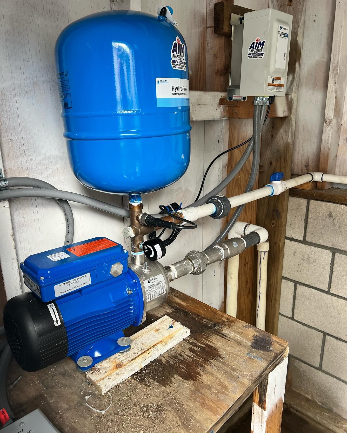 A blue water pump and pressure tank installed on a wooden table against a cinder block wall, with pipes and wiring.