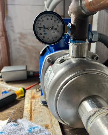 A close-up of a blue electric water pump with an attached pressure gauge, sitting on a surface with a screwdriver nearby.