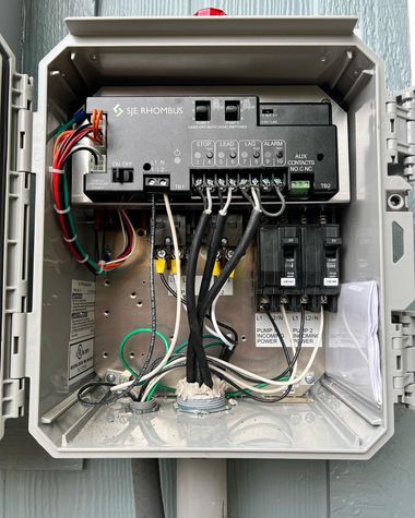 An open electrical control box mounted on a wall, showing a pool controller, circuit breakers, and internal wiring.