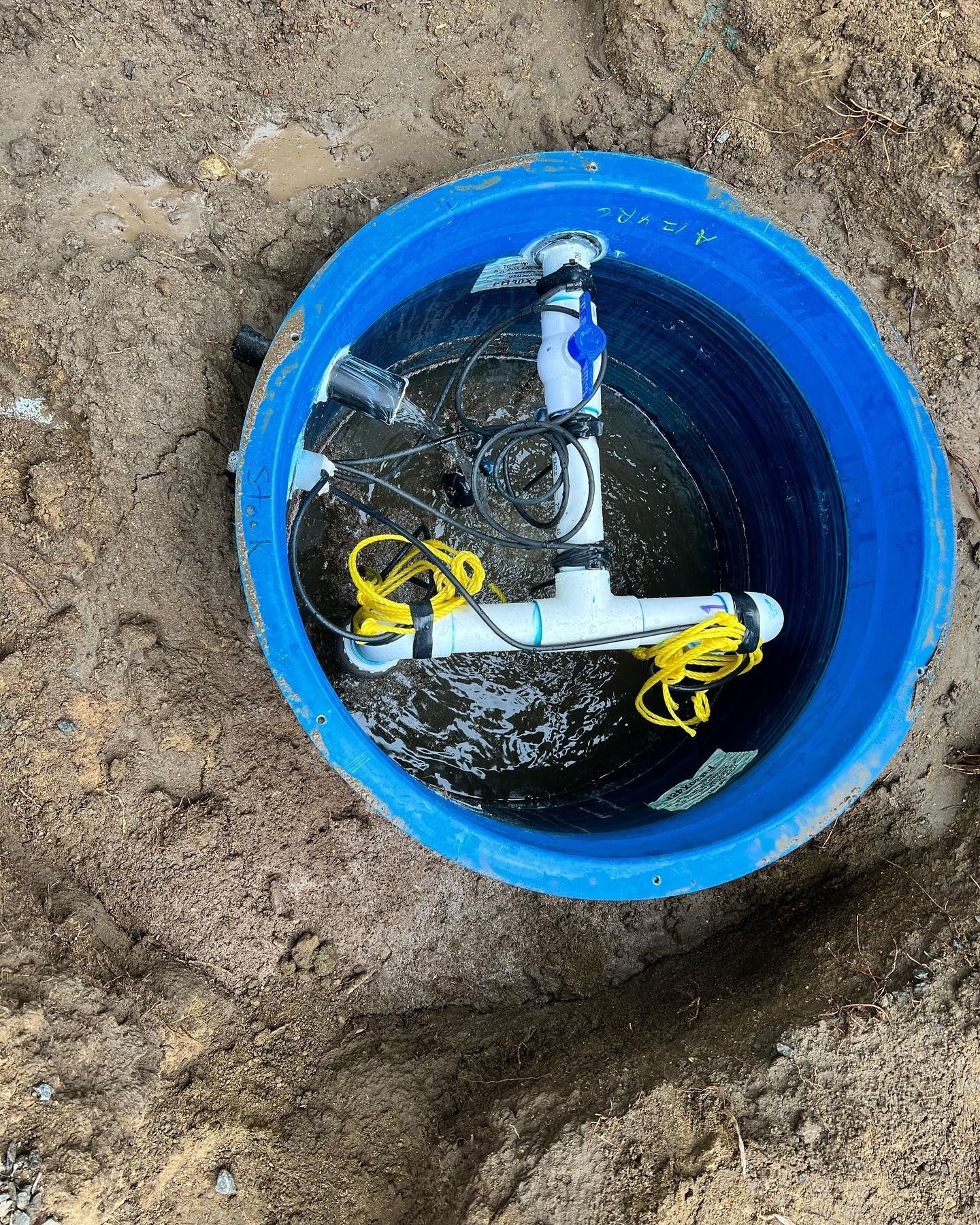 A blue plastic basin in the ground containing white PVC piping, submerged pump components, and coiled yellow cords.