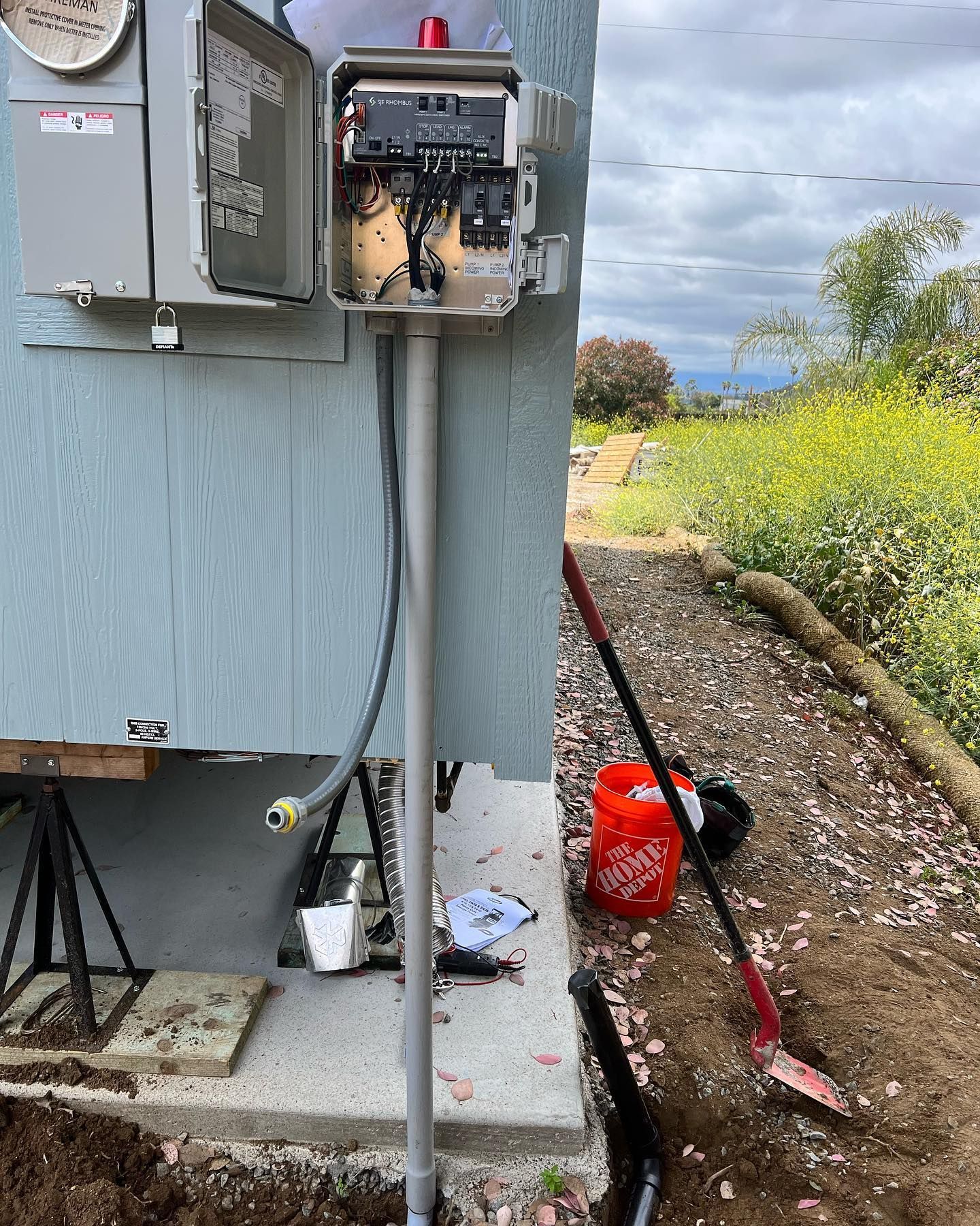 An outdoor electrical box mounted to a siding wall with an exposed, open panel, next to a shovel and bucket on dirt.