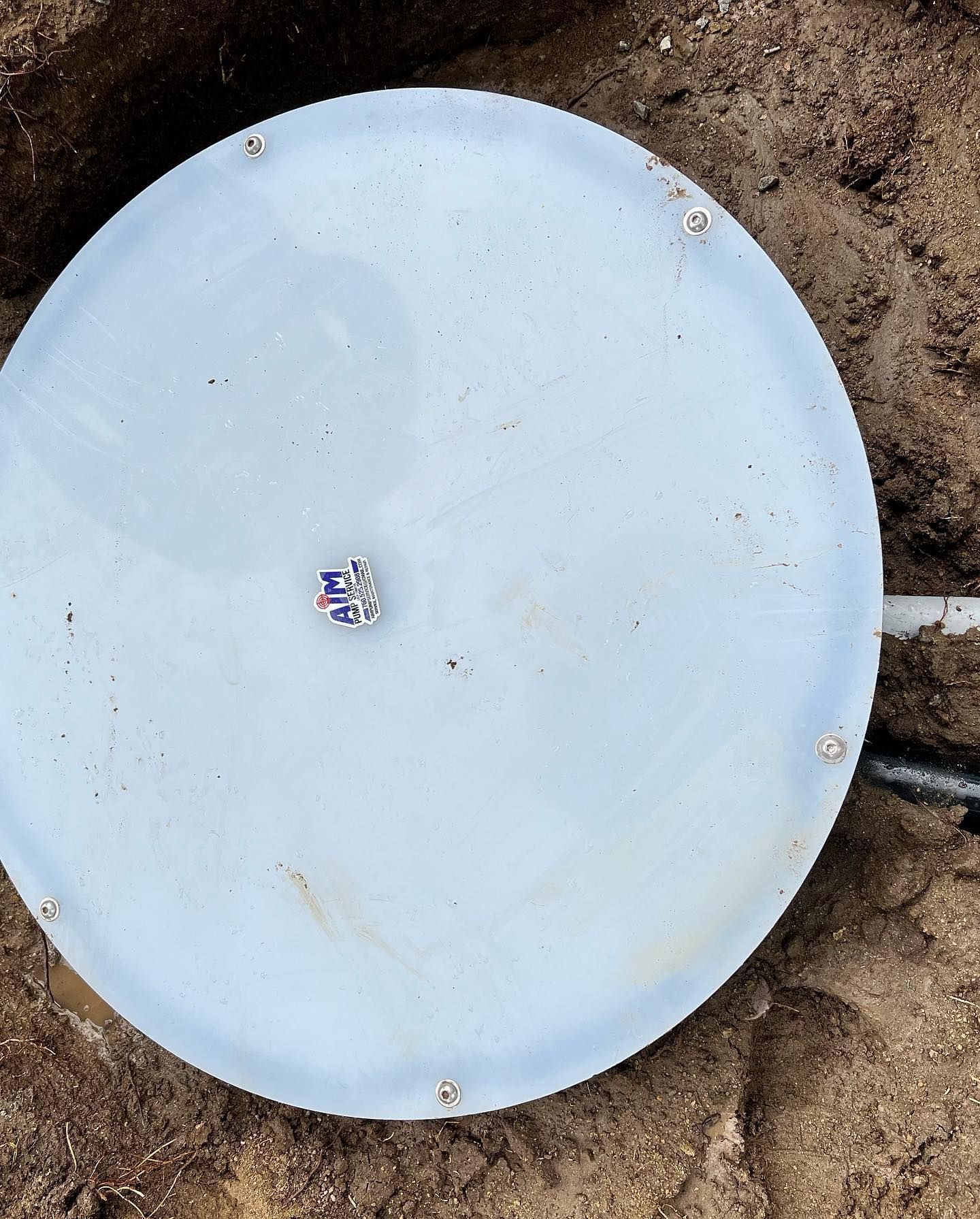 A light blue circular plastic lid secured with screws in a patch of dirt, with a small piece of trash on top.