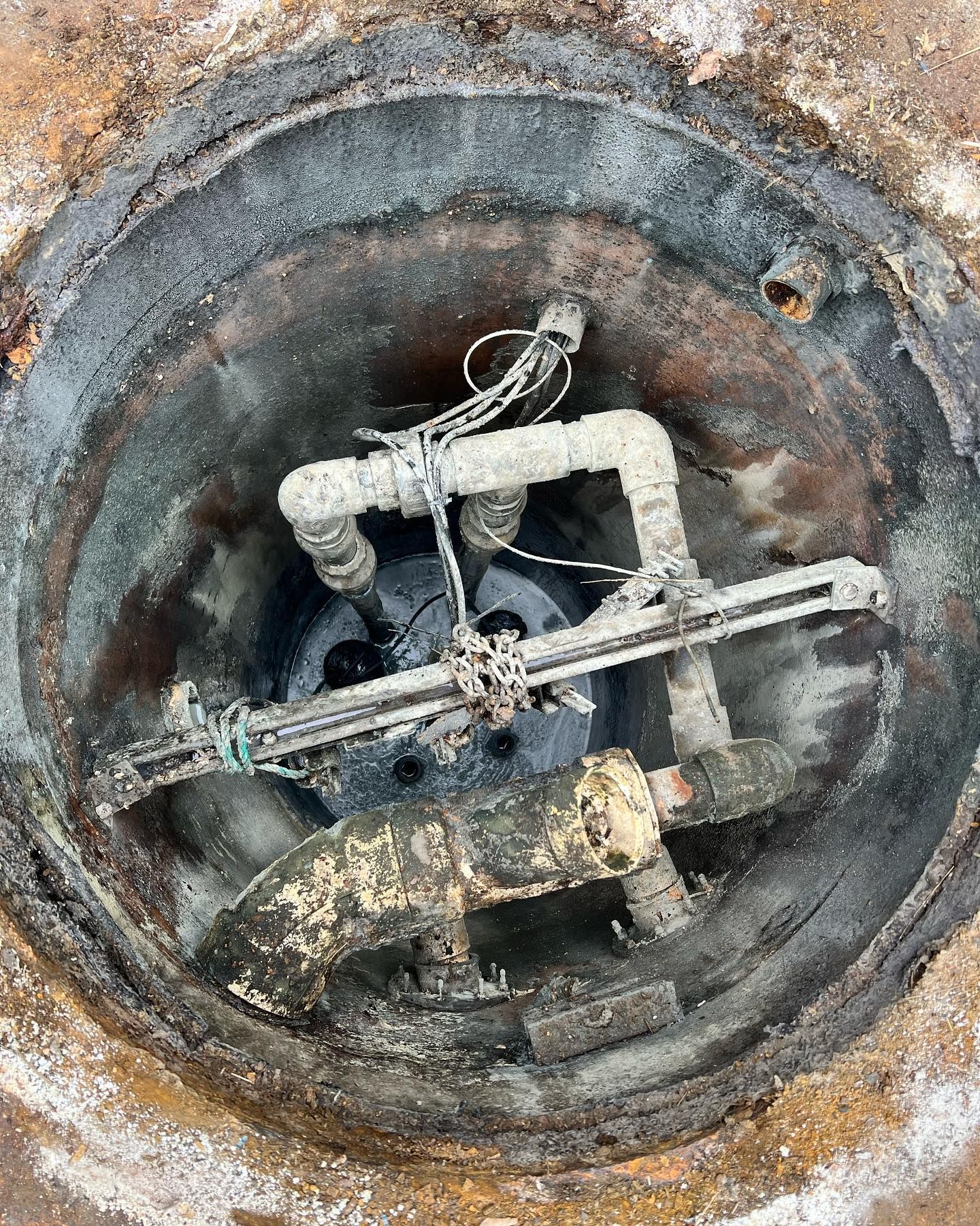 An open underground utility access pit showing weathered pipes, metal support structures, and electrical wiring.
