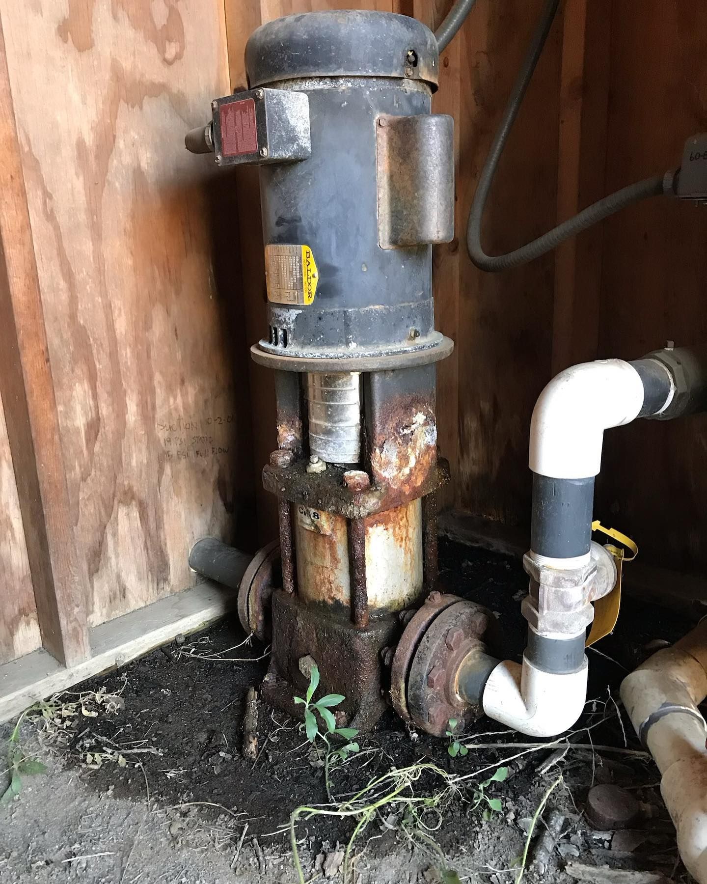 A vertical electric pump with a rusted metal base and motor, connected to white PVC pipes in a shed.