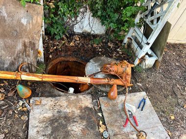 A wooden pole rests across an open, circular septic tank cover outdoors, with tools and brown float switches nearby.