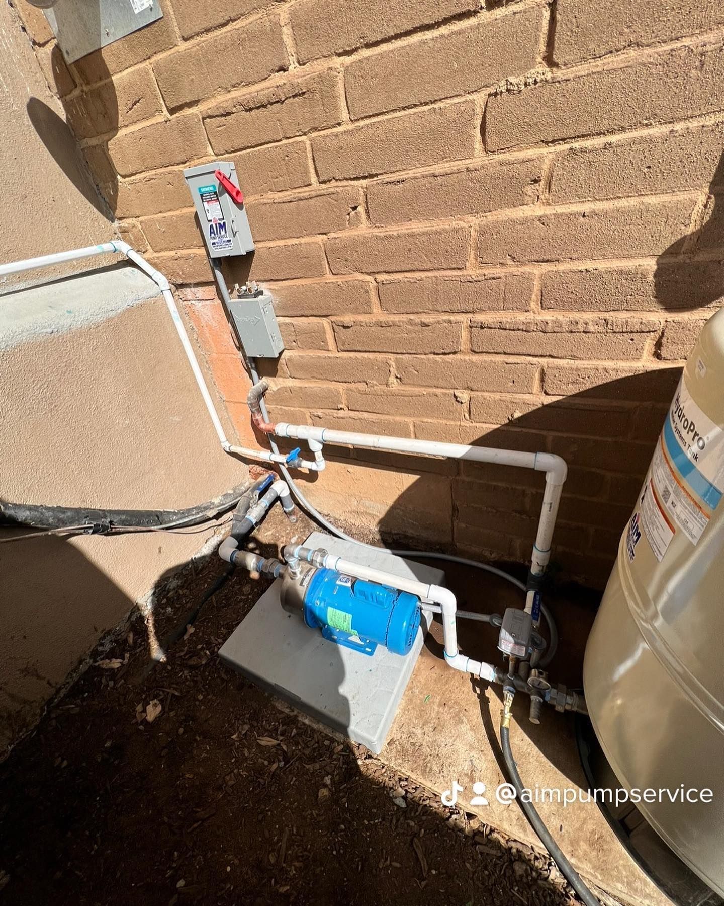 A blue water pump connected to white PVC pipes and a large pressure tank against an outdoor brick wall.