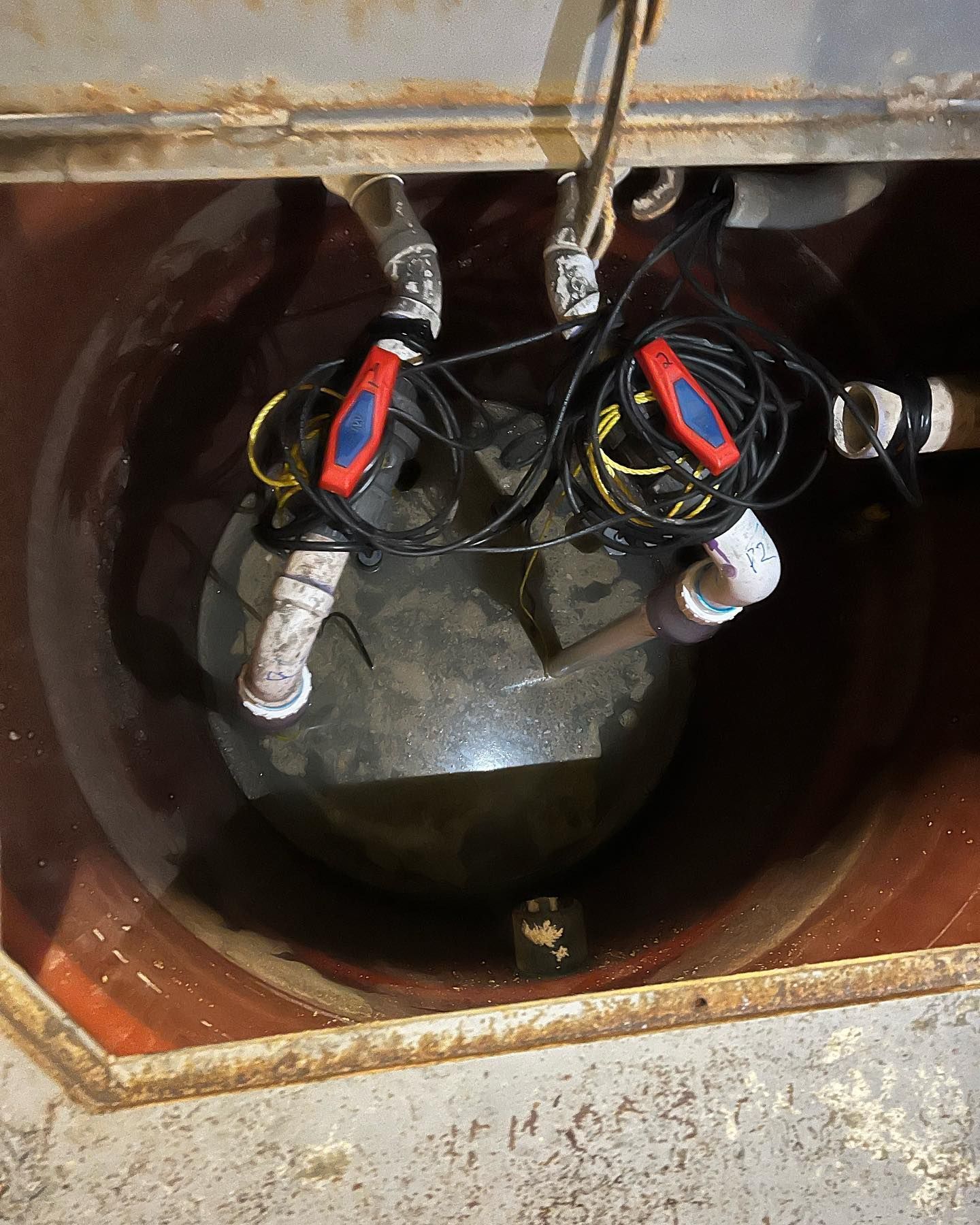 Two sump pumps sit inside a concrete basin, featuring red-handled valves, white piping, and black electrical cords.