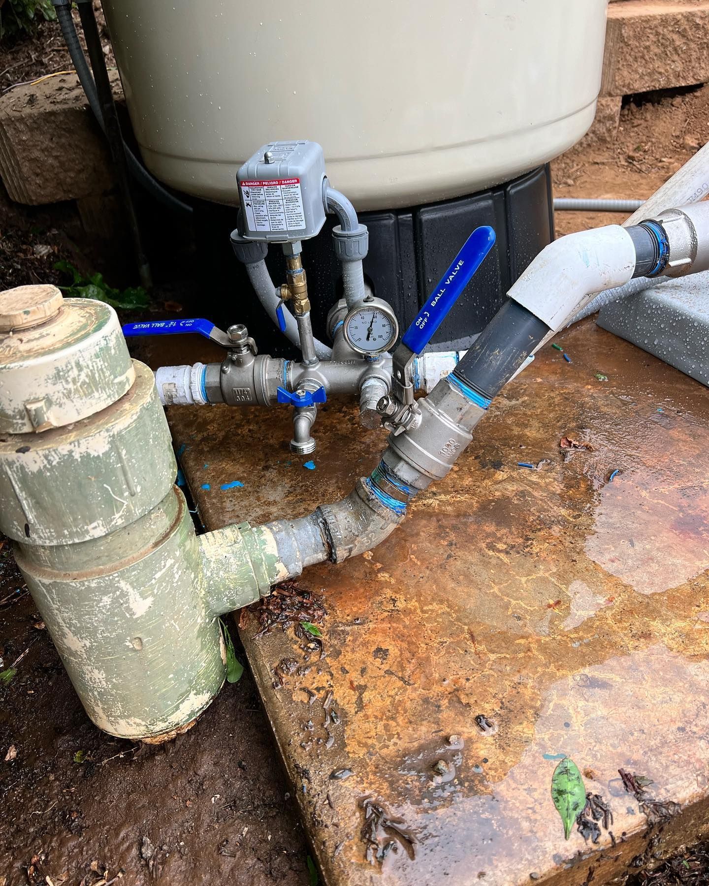 Outdoor pool filtration system showing pipes, blue ball valves, a pressure gauge, and a pump housing on a stone patio.