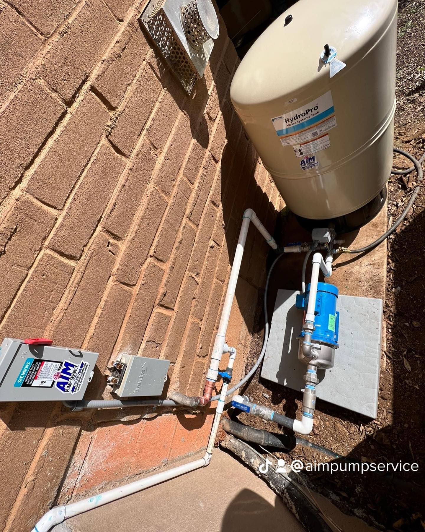 A beige well pressure tank and a blue pump installed on a concrete slab next to a brick wall.