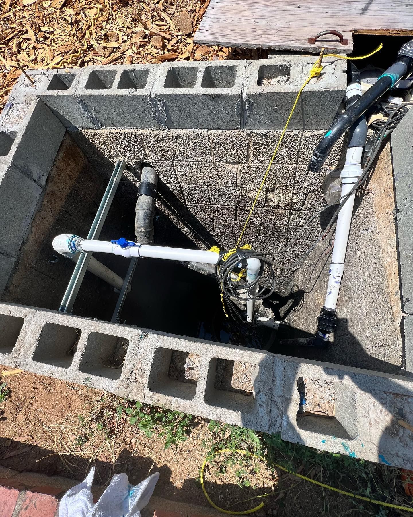 A pump installation inside a rectangular cinder block well, featuring white PVC pipes, black tubing, and a yellow rope.
