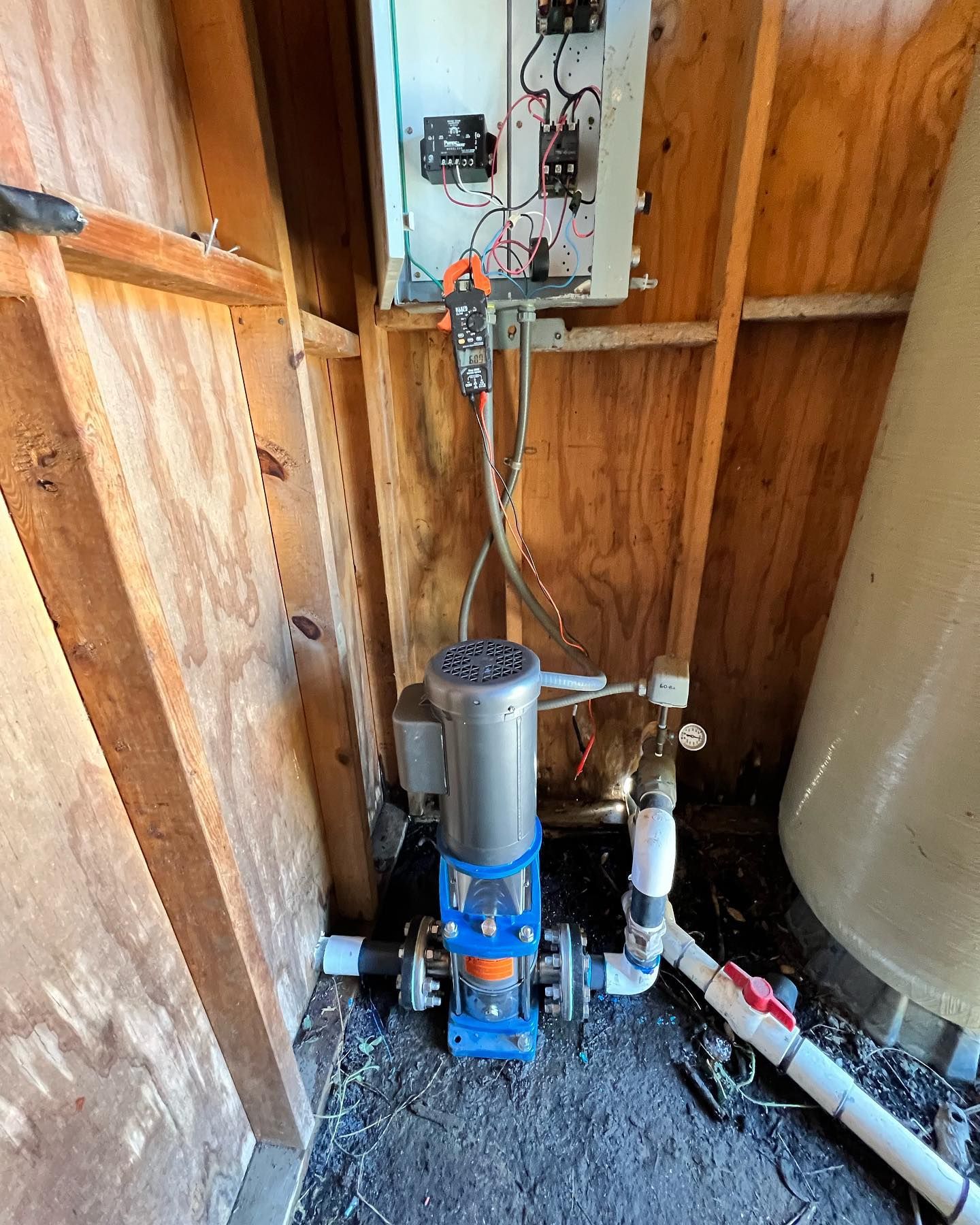 A blue vertical water pump connected to pipes inside a wooden enclosure, with electrical equipment mounted on the wall.