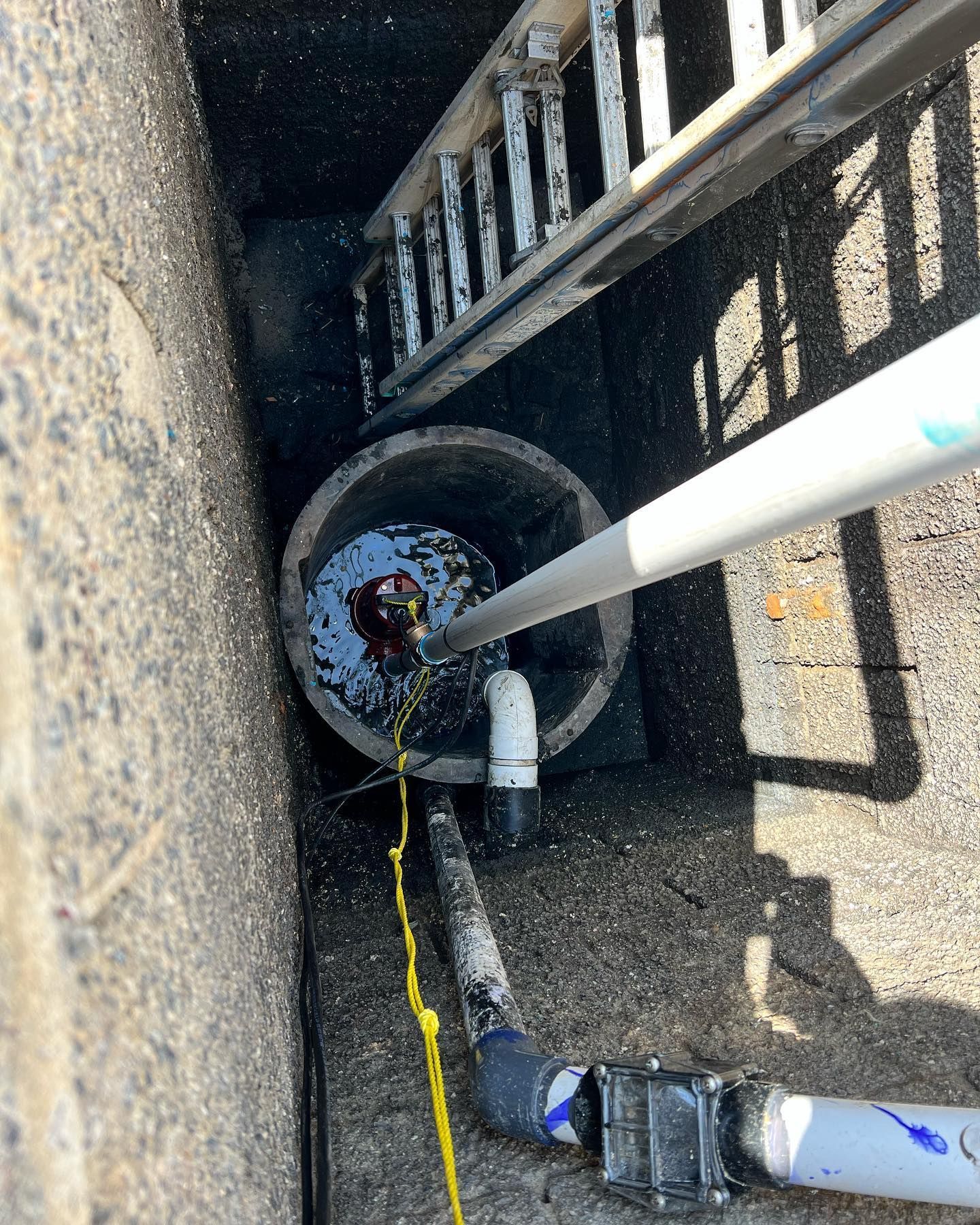 A submersible pump sits in a water-filled basin within a concrete shaft, connected to white pipes and a yellow cable.