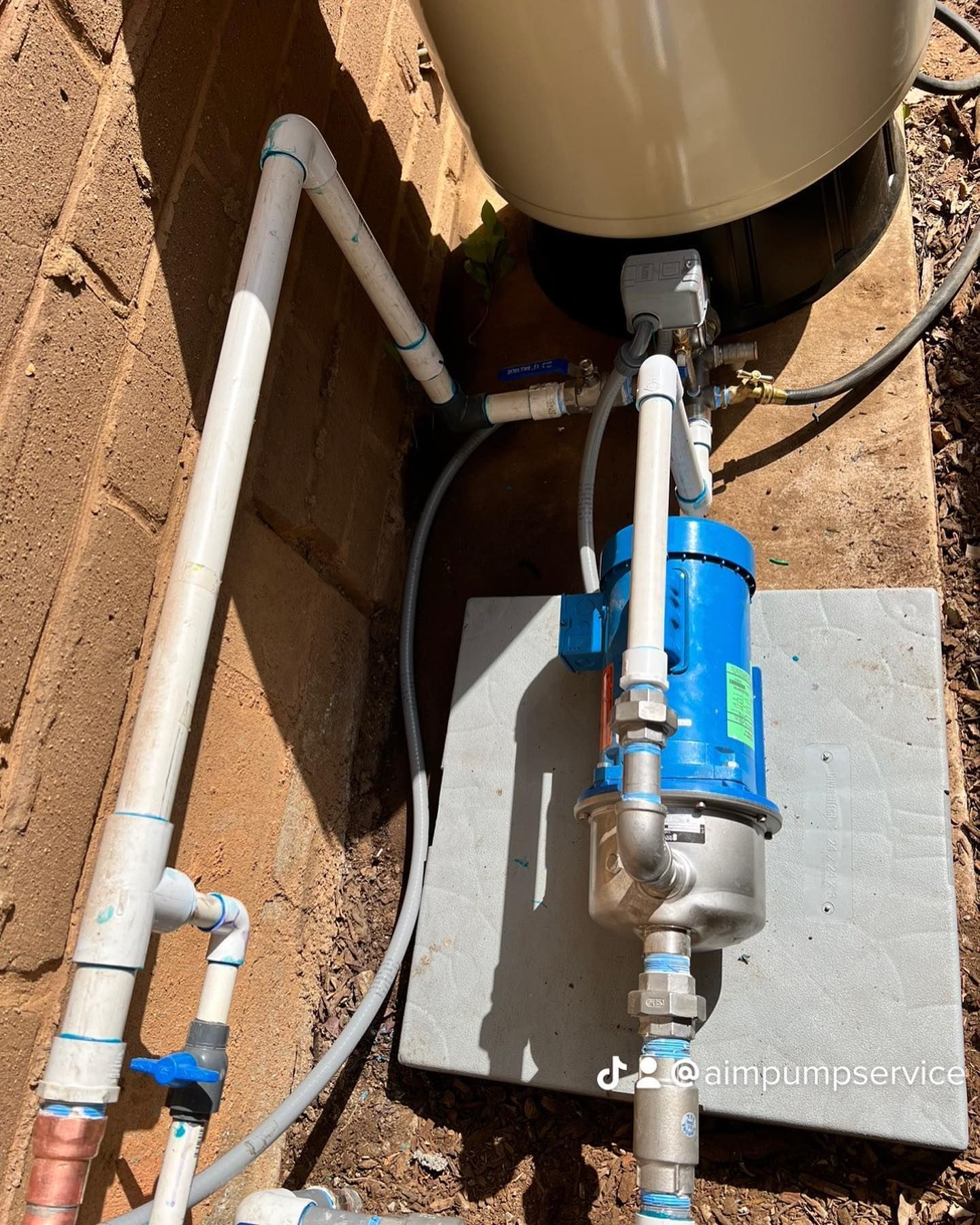 A blue electric pump and white PVC piping system connected to a large water filtration tank on a concrete pad.