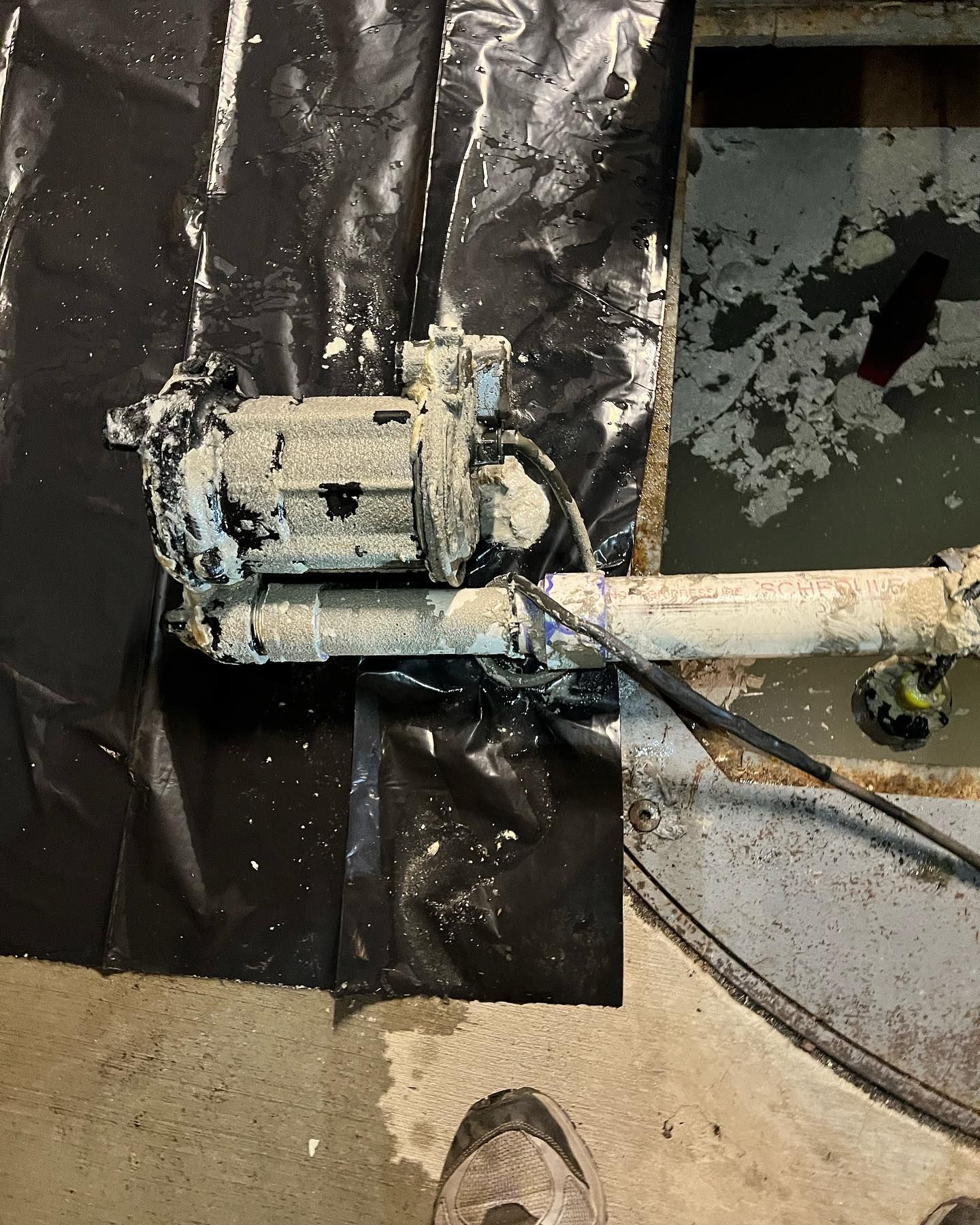 A dusty sump pump motor with a black power cord resting on a black plastic sheet near an open basement floor drain.
