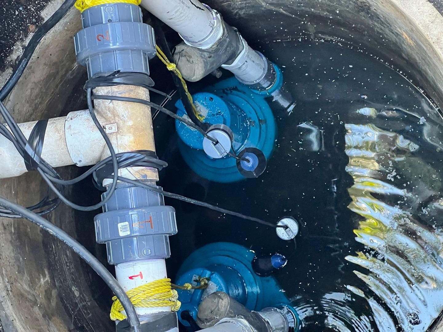 Two blue sump pumps submerged in a dark utility basin, connected to white PVC plumbing pipes.