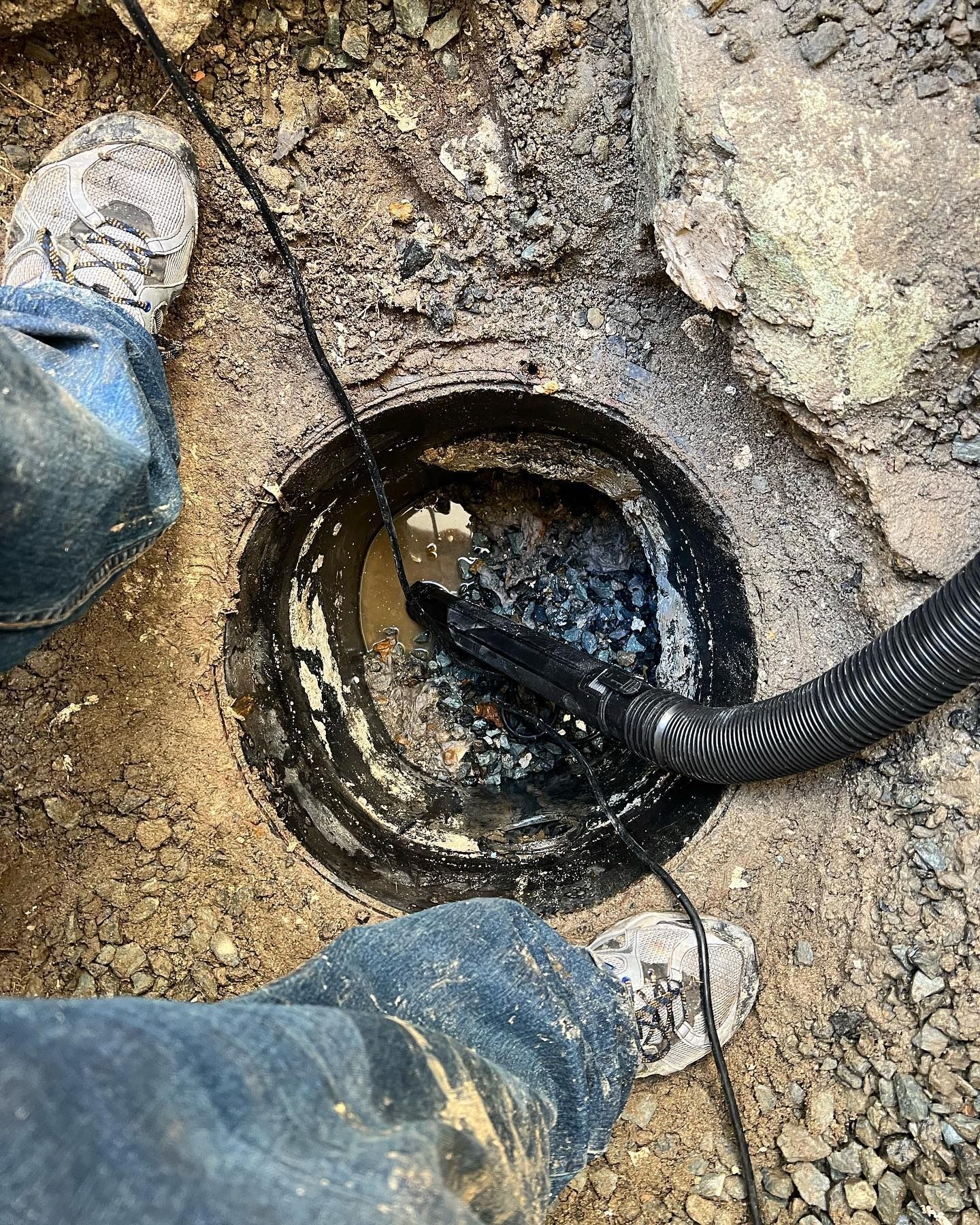 A person uses a vacuum hose to remove dark, wet debris from a circular drainage basin in the ground.