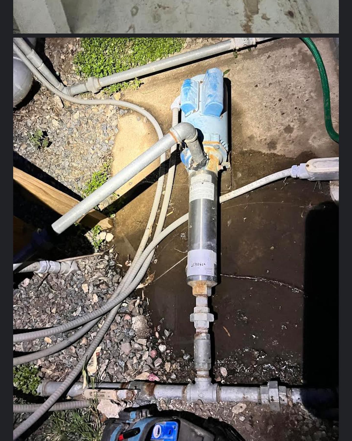 A pump assembly with gray piping and a blue motor situated on a patch of ground, surrounded by wiring and gravel.