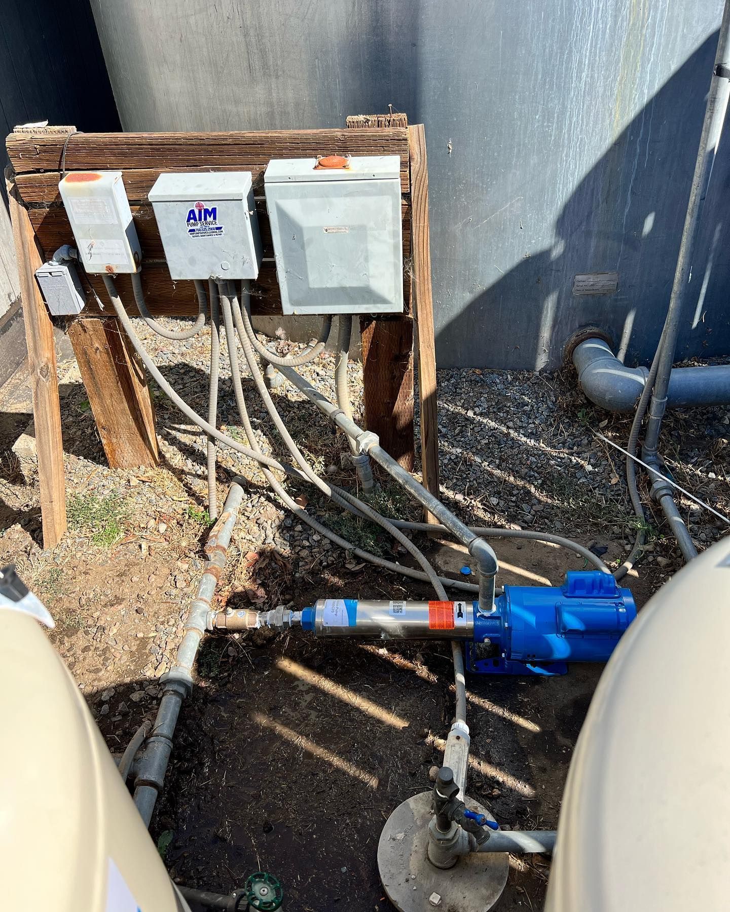 A blue water pump and electrical control boxes mounted on a wooden frame outdoors near large tanks.