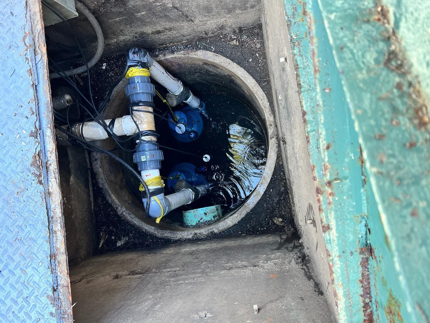 Two industrial water pumps submerged in a round concrete basin, connected by white PVC pipes with blue valves.