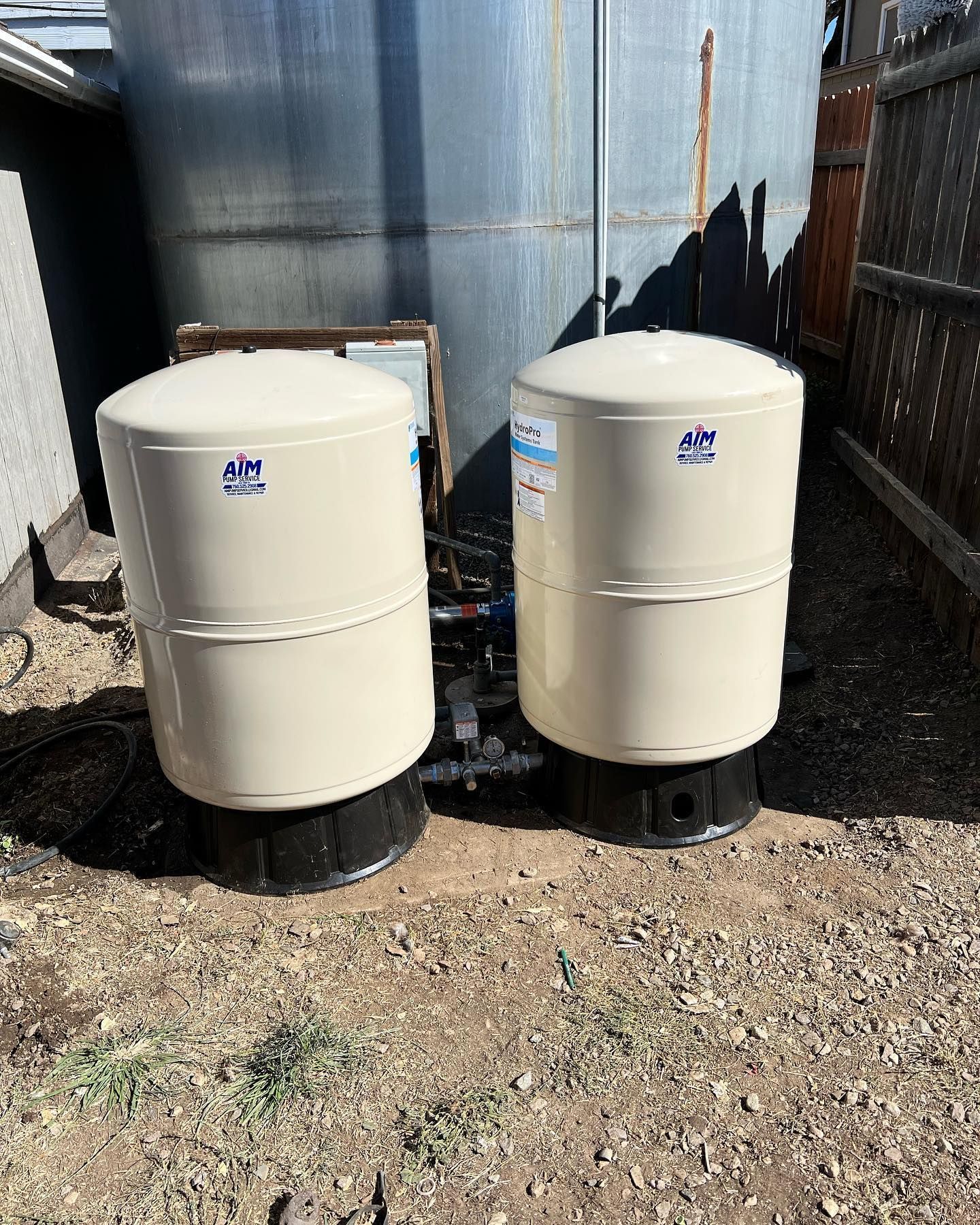 Two light-tan water pressure tanks with black bases sit side-by-side on gravel in front of a large metal storage tank.