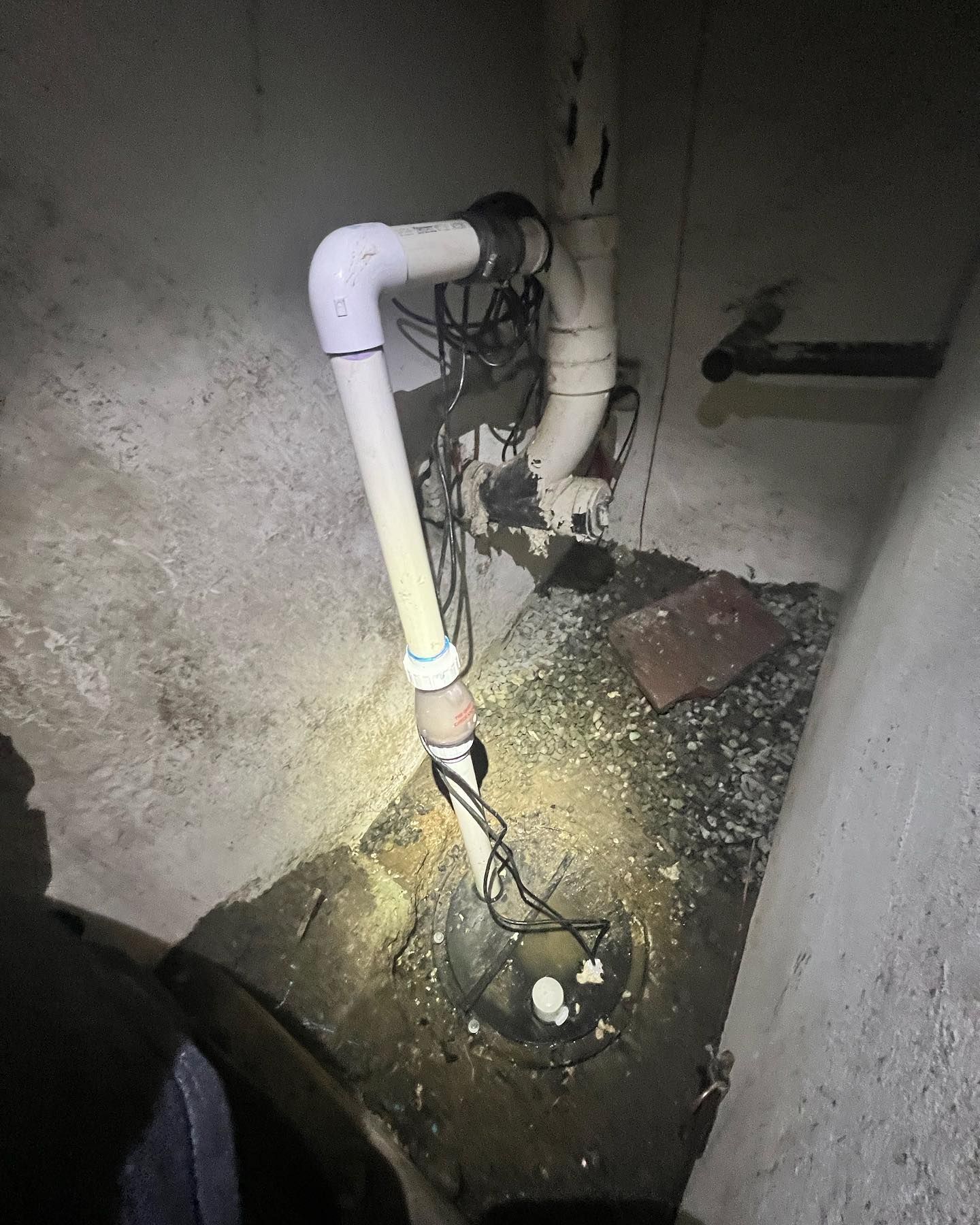 A white sump pump pipe extends from a basement floor into a vertical discharge pipe, with electrical cords visible.