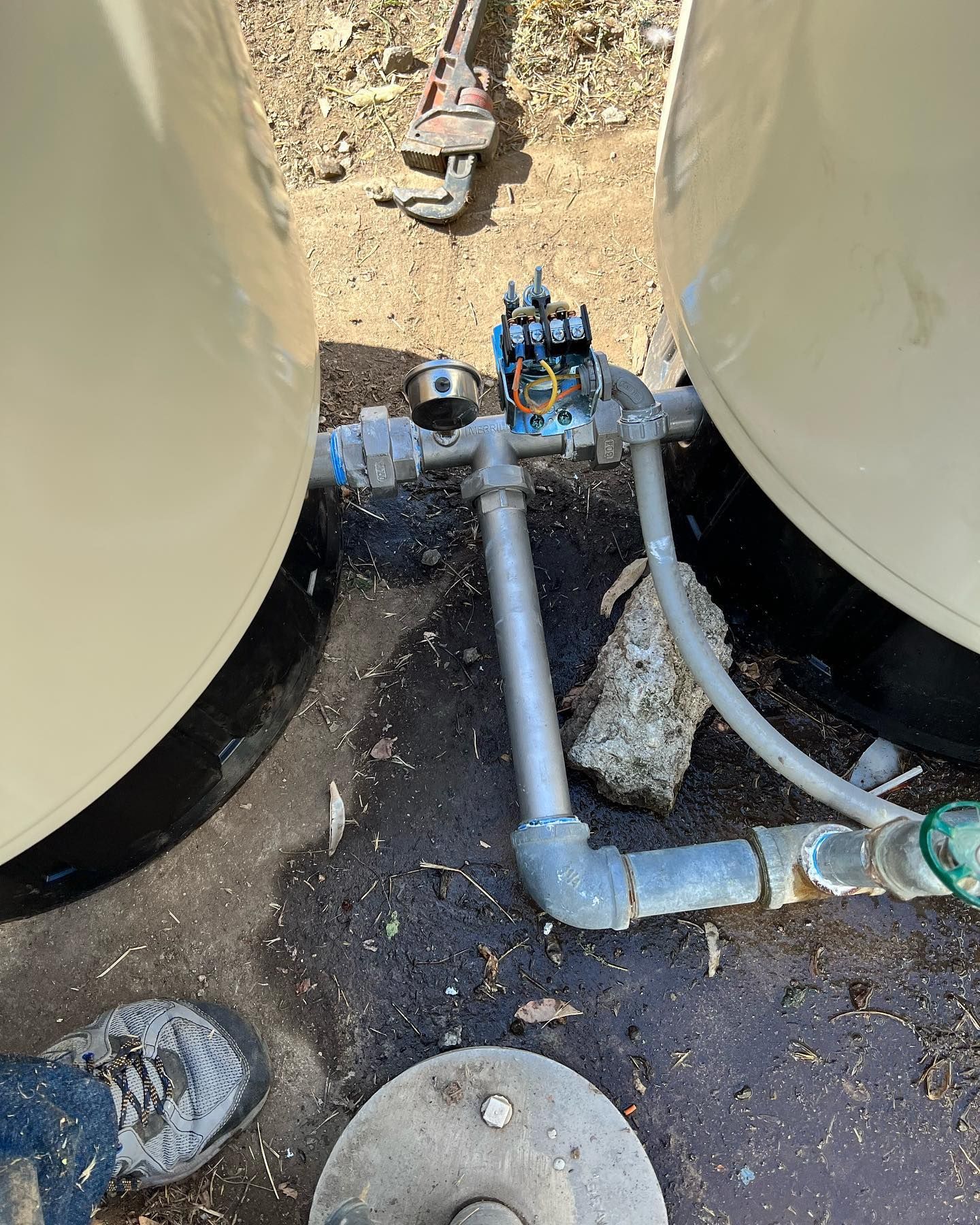 Two large beige tanks connected by galvanized pipes, a pressure gauge, and a pressure switch on a concrete ground surface.