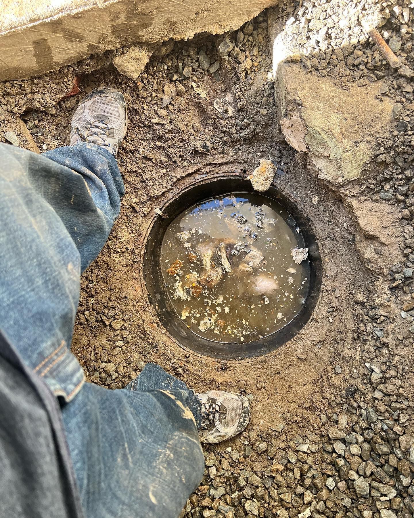 A high-angle view of a person standing near an open, dirt-filled ground hole containing murky, debris-filled water.