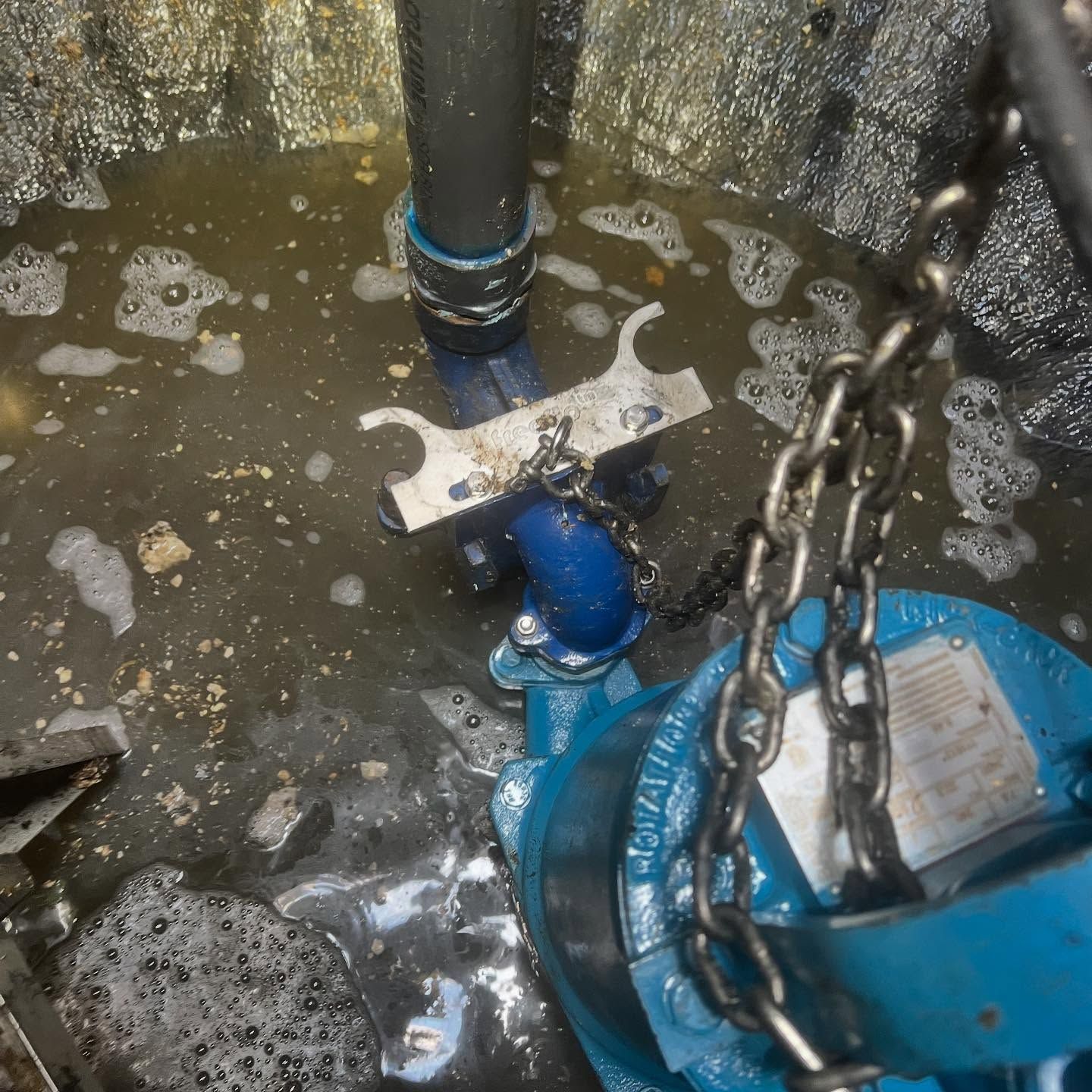 A blue submersible pump sits in murky water, connected to a vertical pipe and a metal lift chain inside a basin.
