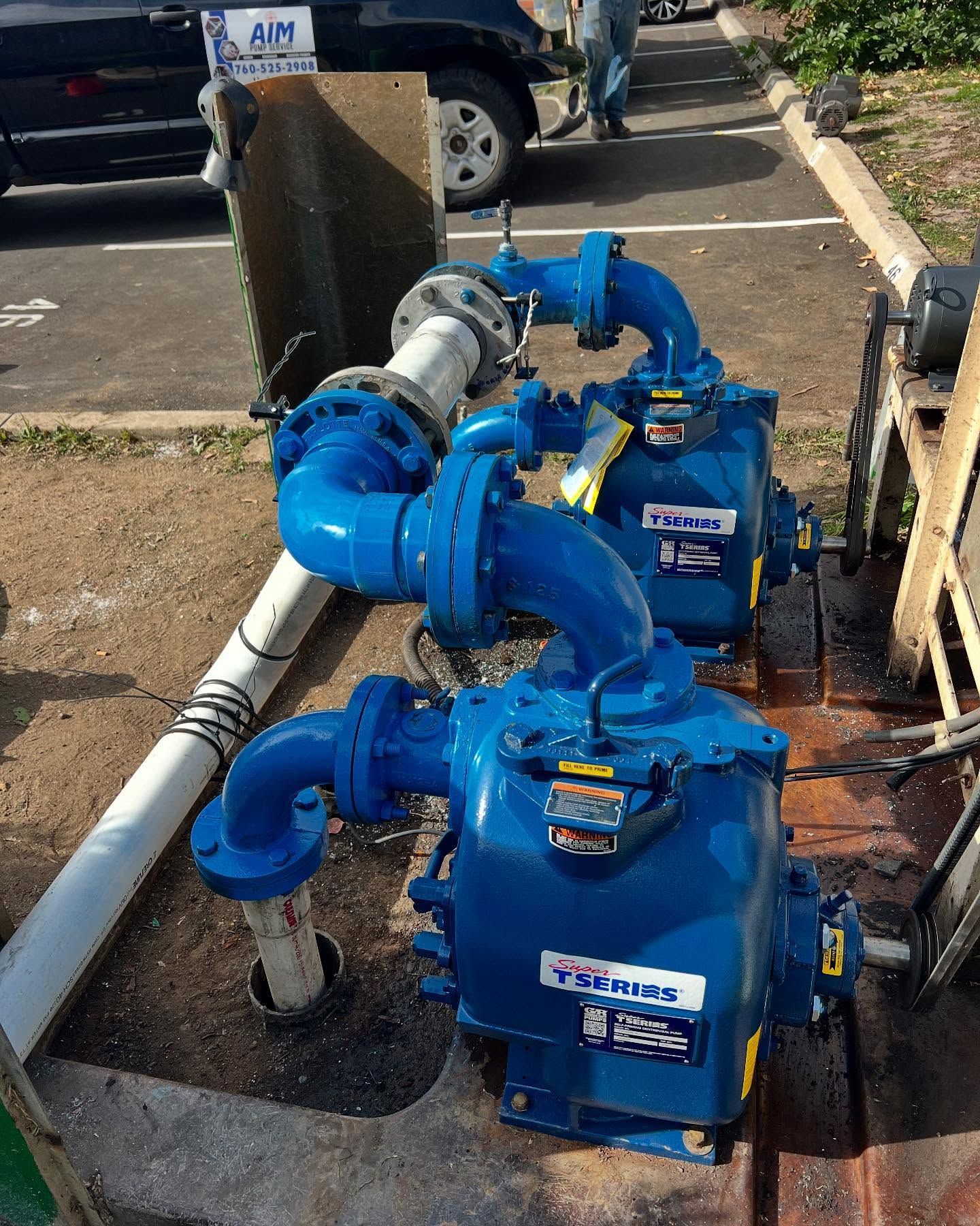 Two blue industrial pumps connected by pipes on a dirt lot, with a vehicle parked in the background.
