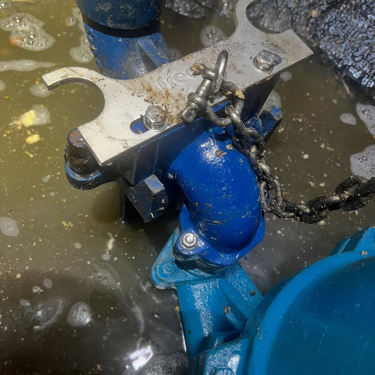 A submerged blue industrial pump connected by a metal bracket and chain in murky, foamy water.