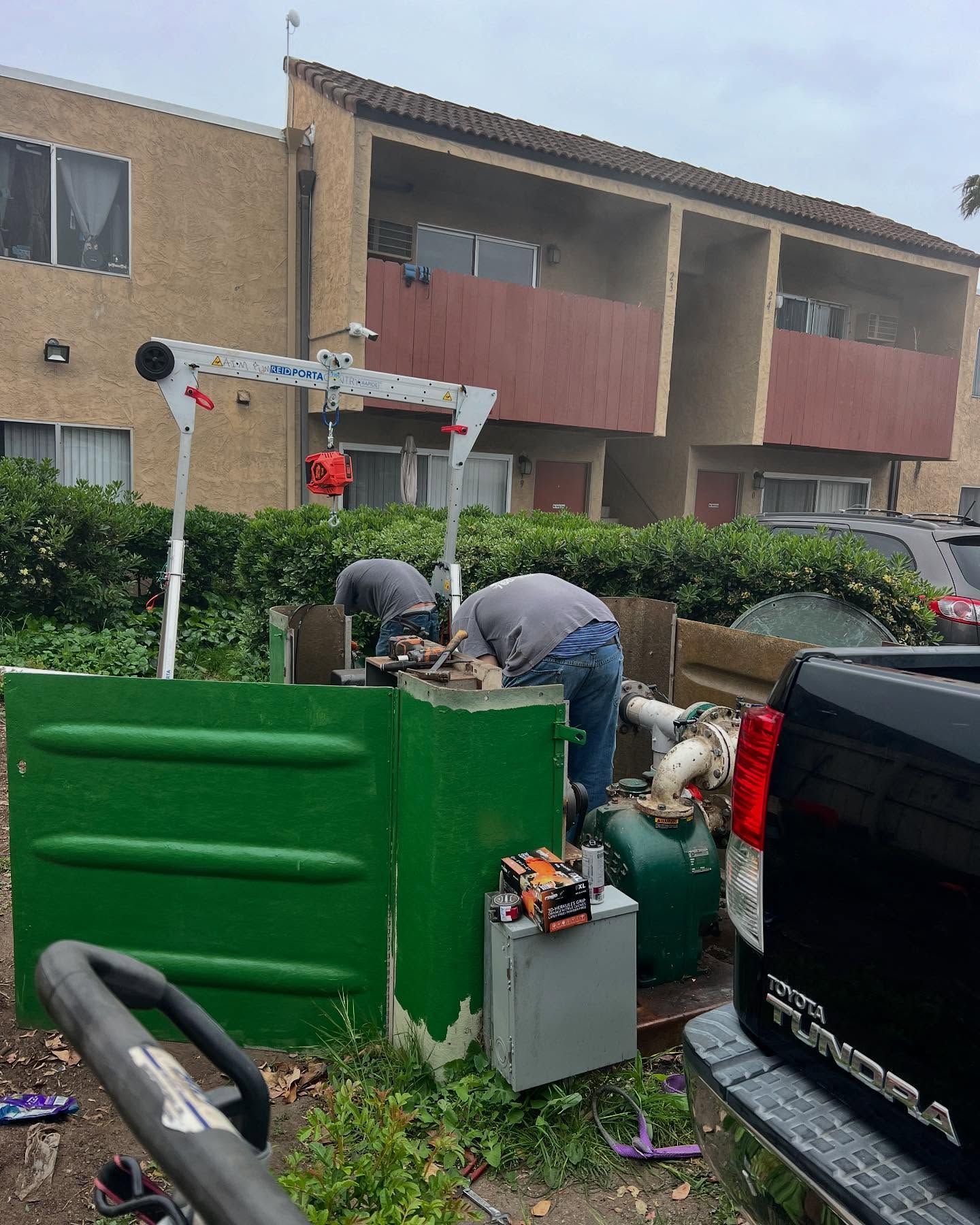 Two workers in grey shirts repair mechanical equipment at an apartment building using a portable tripod hoist.