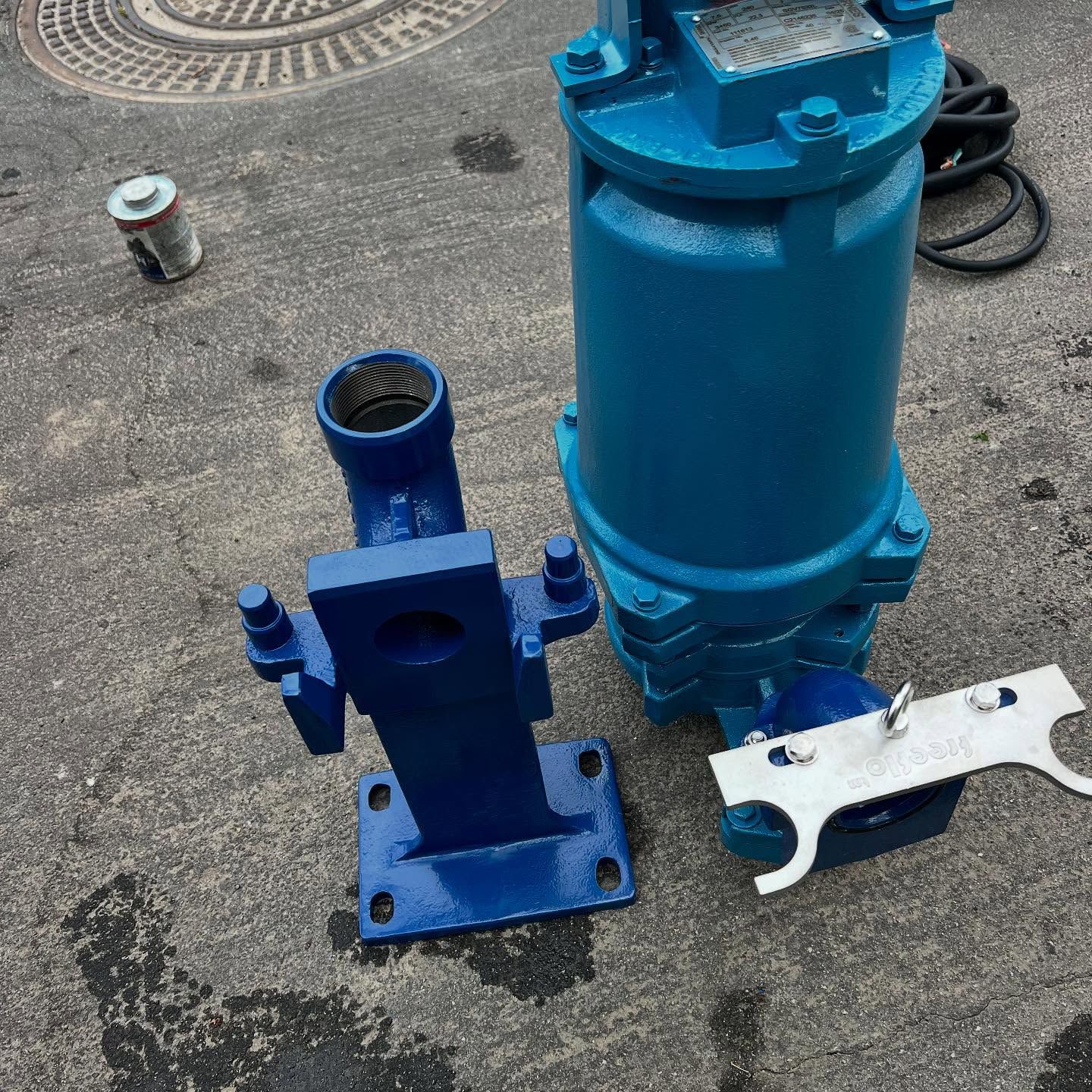 A blue submersible pump lies on asphalt next to its mounting bracket and a silver metal plate.