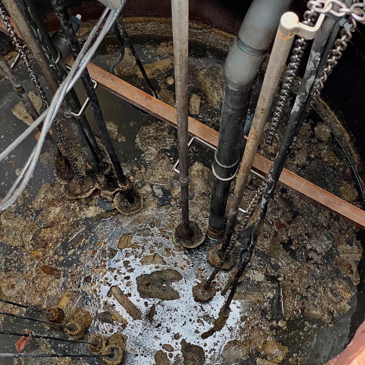 A high-angle view into a dark circular tank filled with murky water, sludge, and various industrial pipes and chains.
