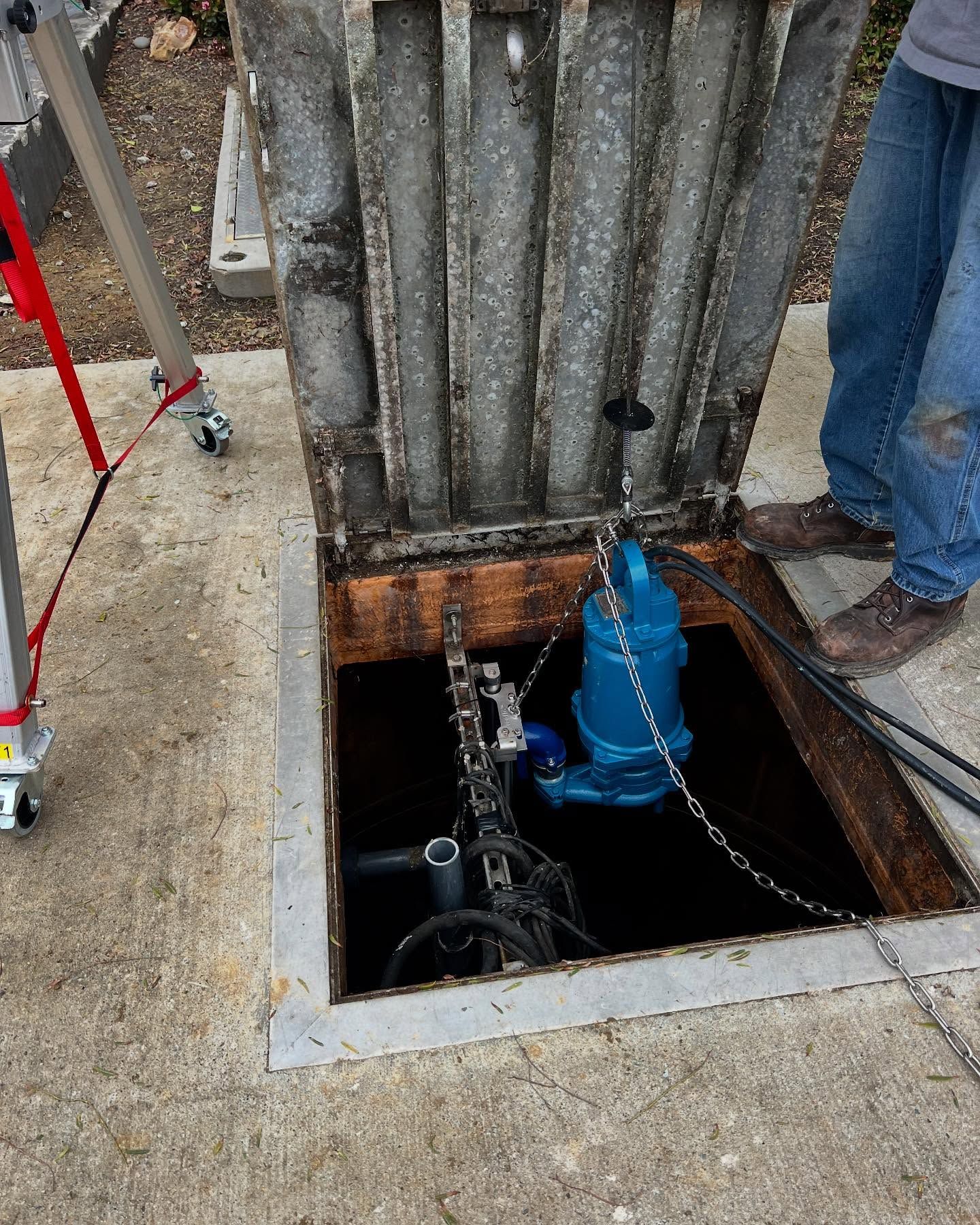 A worker operates a blue industrial pump lowered into an open underground concrete maintenance pit.