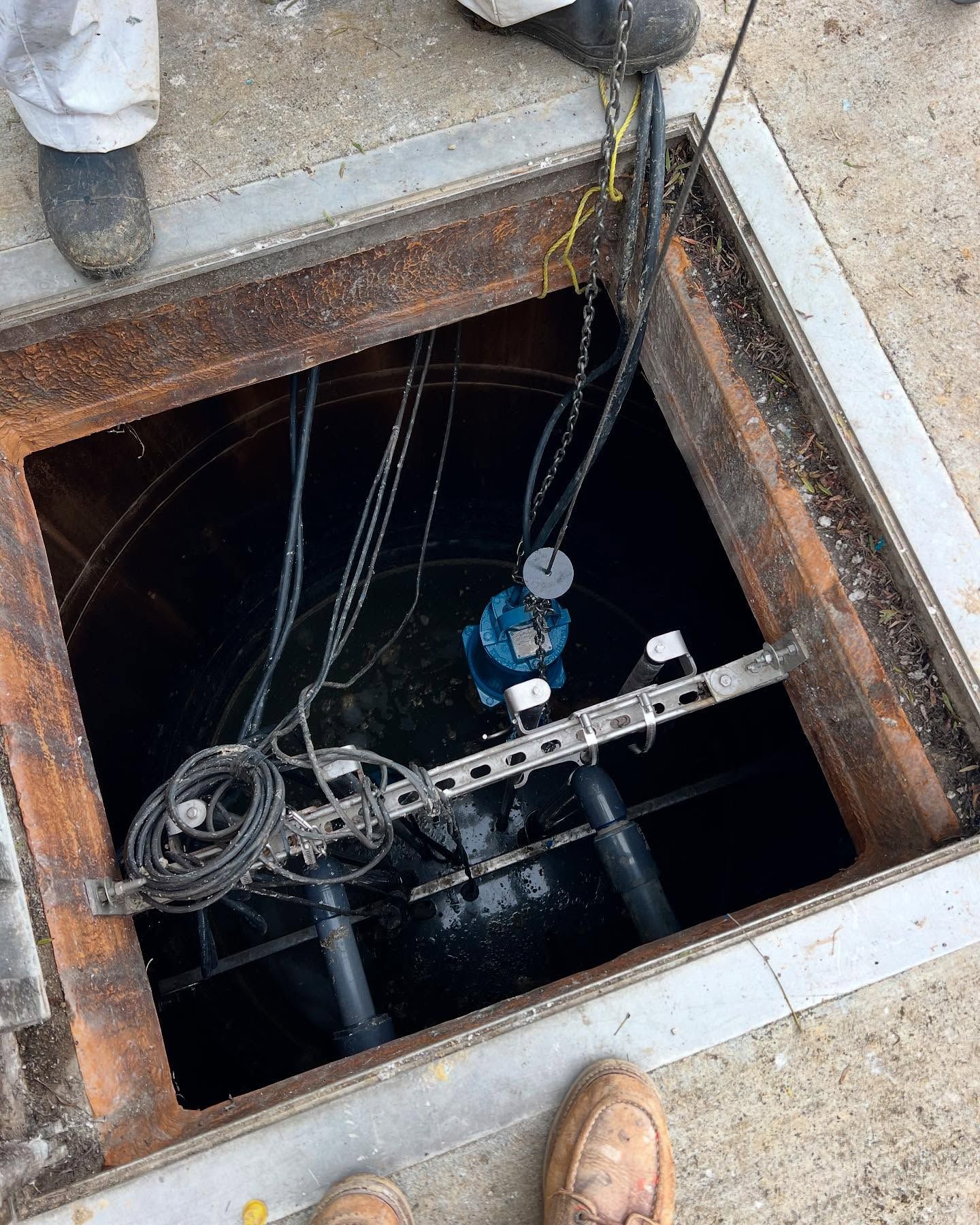 A view into an open industrial manhole revealing a submerged blue pump, wiring, and metal support struts.