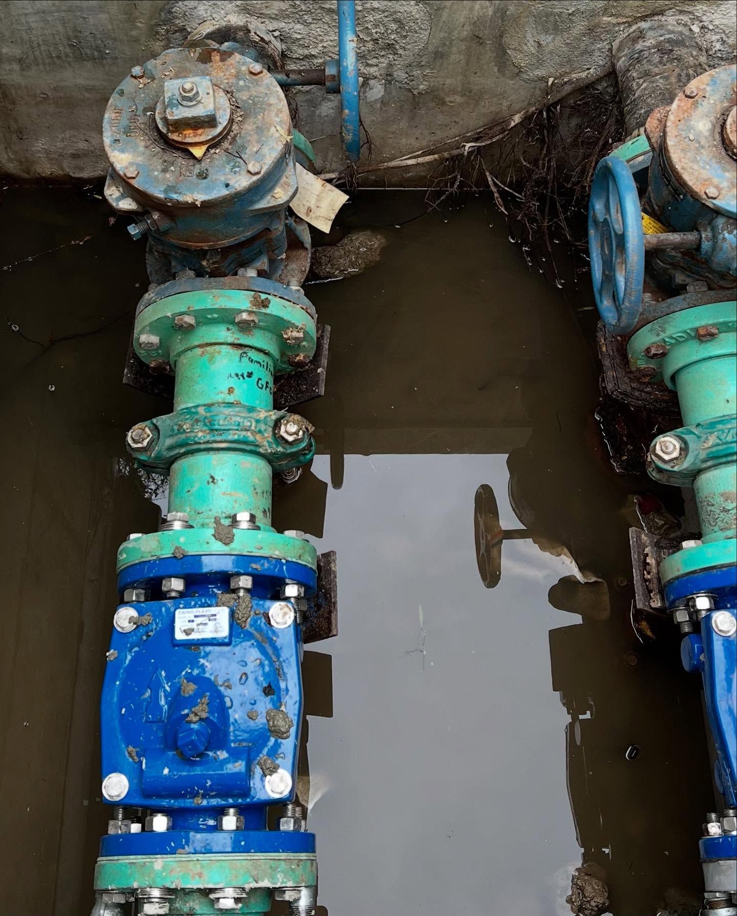 Two industrial green and blue water valves installed in a narrow, partially flooded concrete trench.