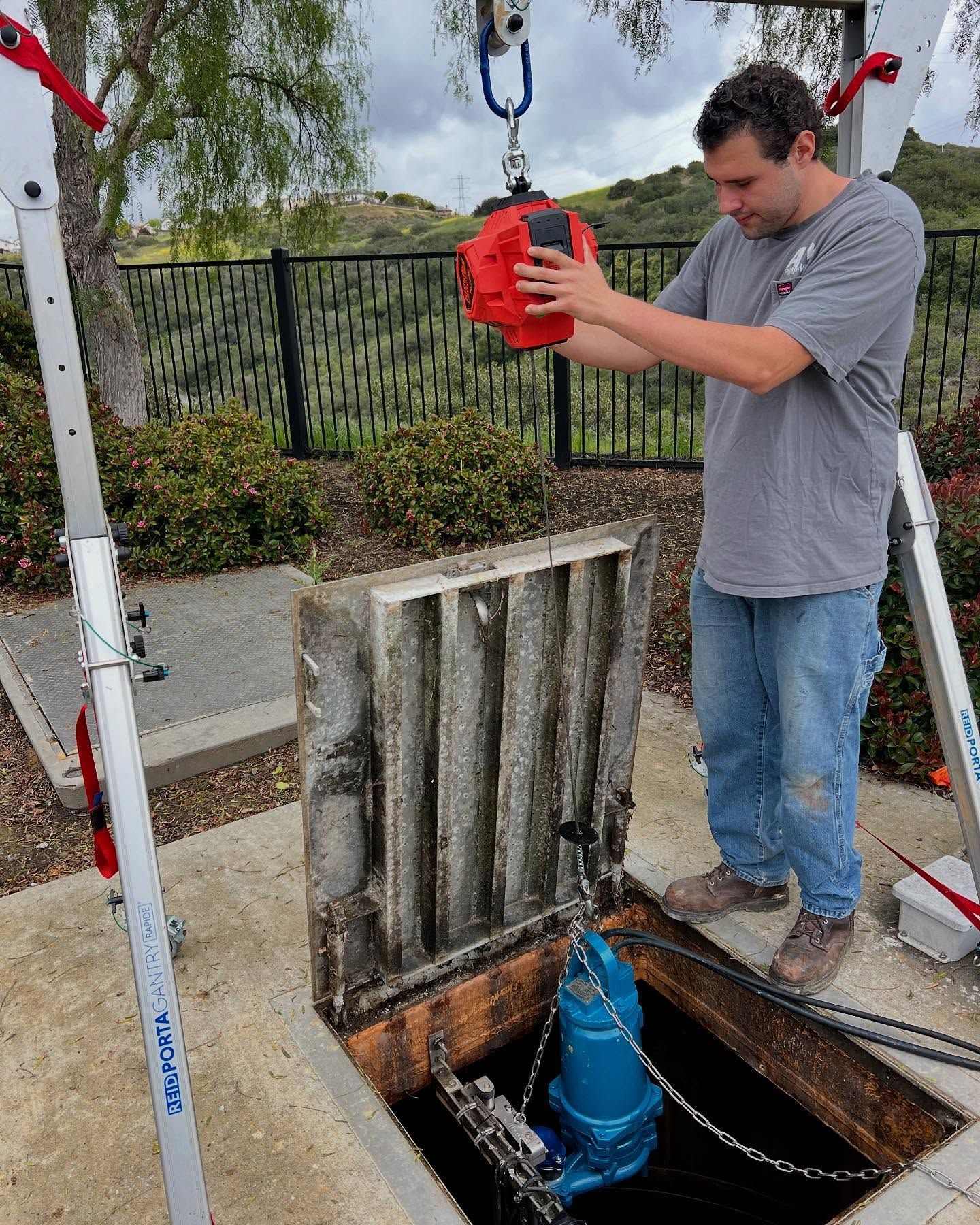 A person uses a portable tripod hoist to lift a blue submersible pump from an open utility vault.