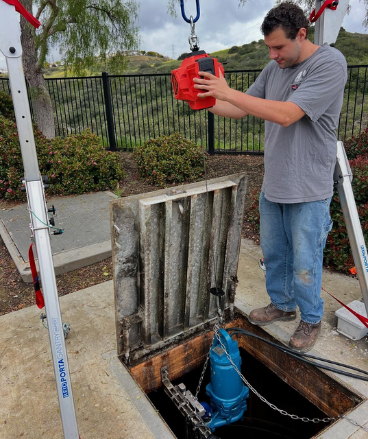 A person in a gray shirt uses a hoist to lift a blue submersible pump from an open concrete maintenance vault.