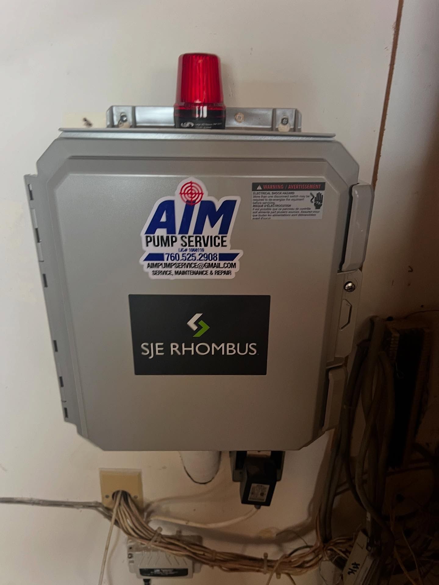 A grey SJE Rhombus pump control box mounted on a wall with a red warning light on top and wiring below.