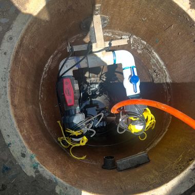 Top-down view of two water pumps inside a round, concrete well, connected by white PVC pipes with blue and red valves.