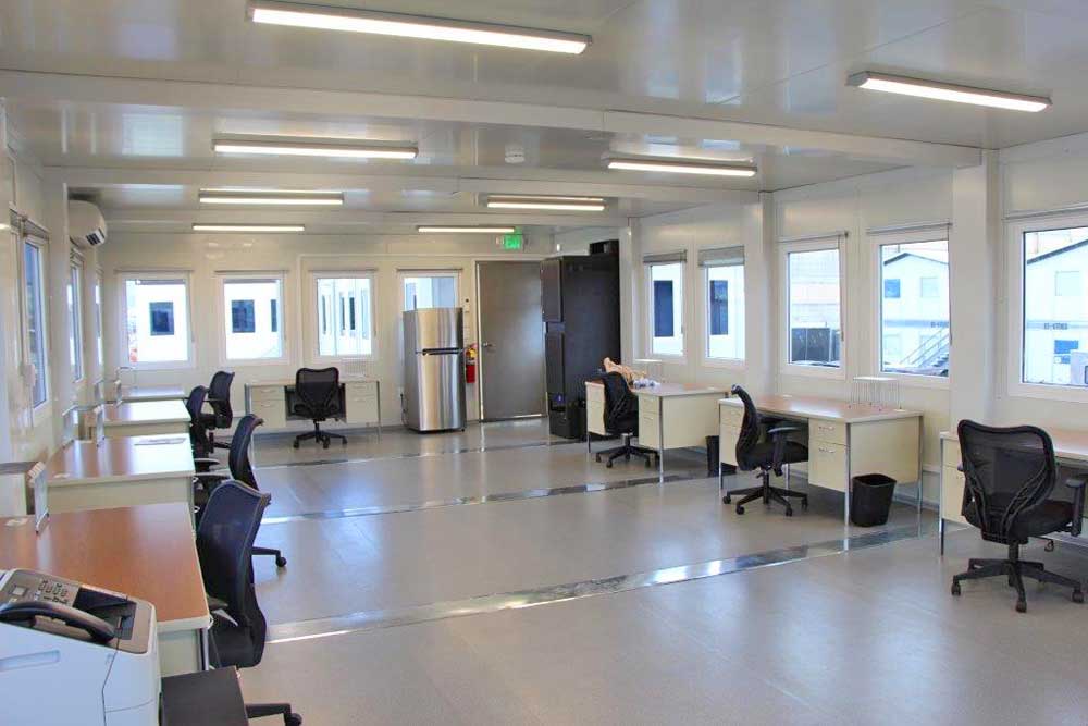 A large office with lots of desks and chairs and a refrigerator.