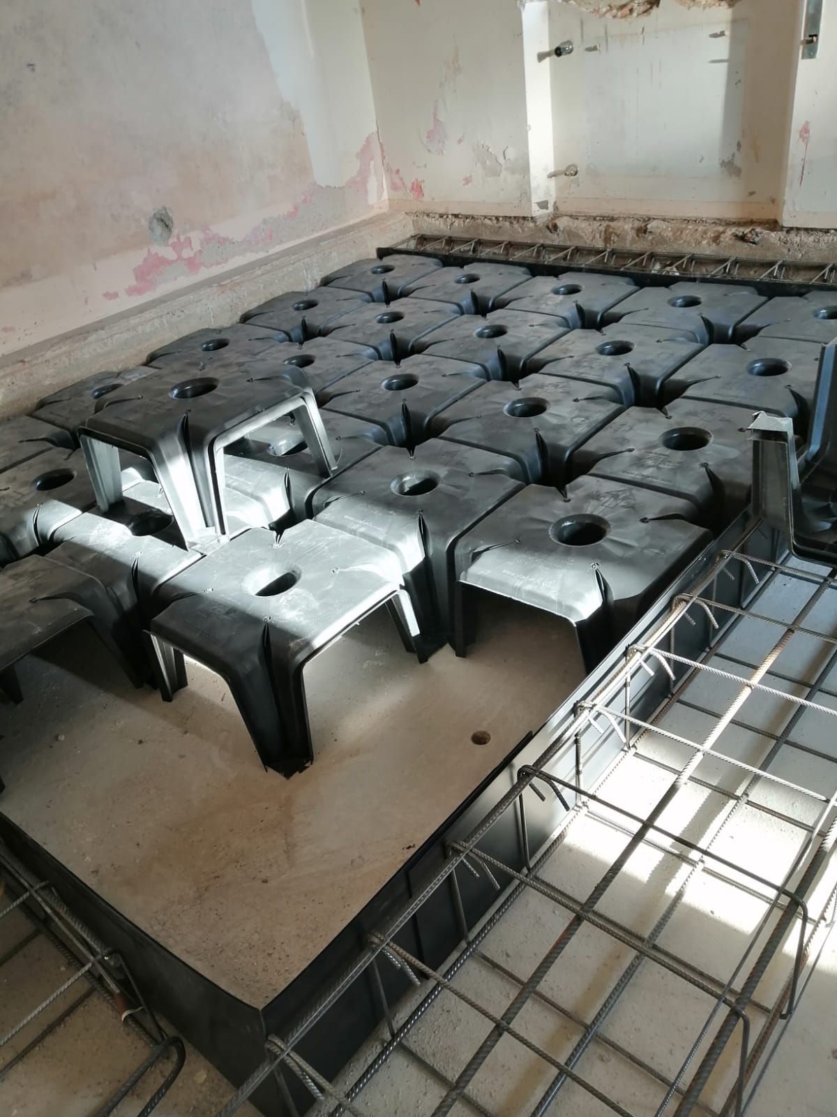 Interno di un cantiere edile con elementi di riempimento in plastica disposti sul sottofondo accanto all'intelaiatura in barre metalliche per una soletta in cemento.