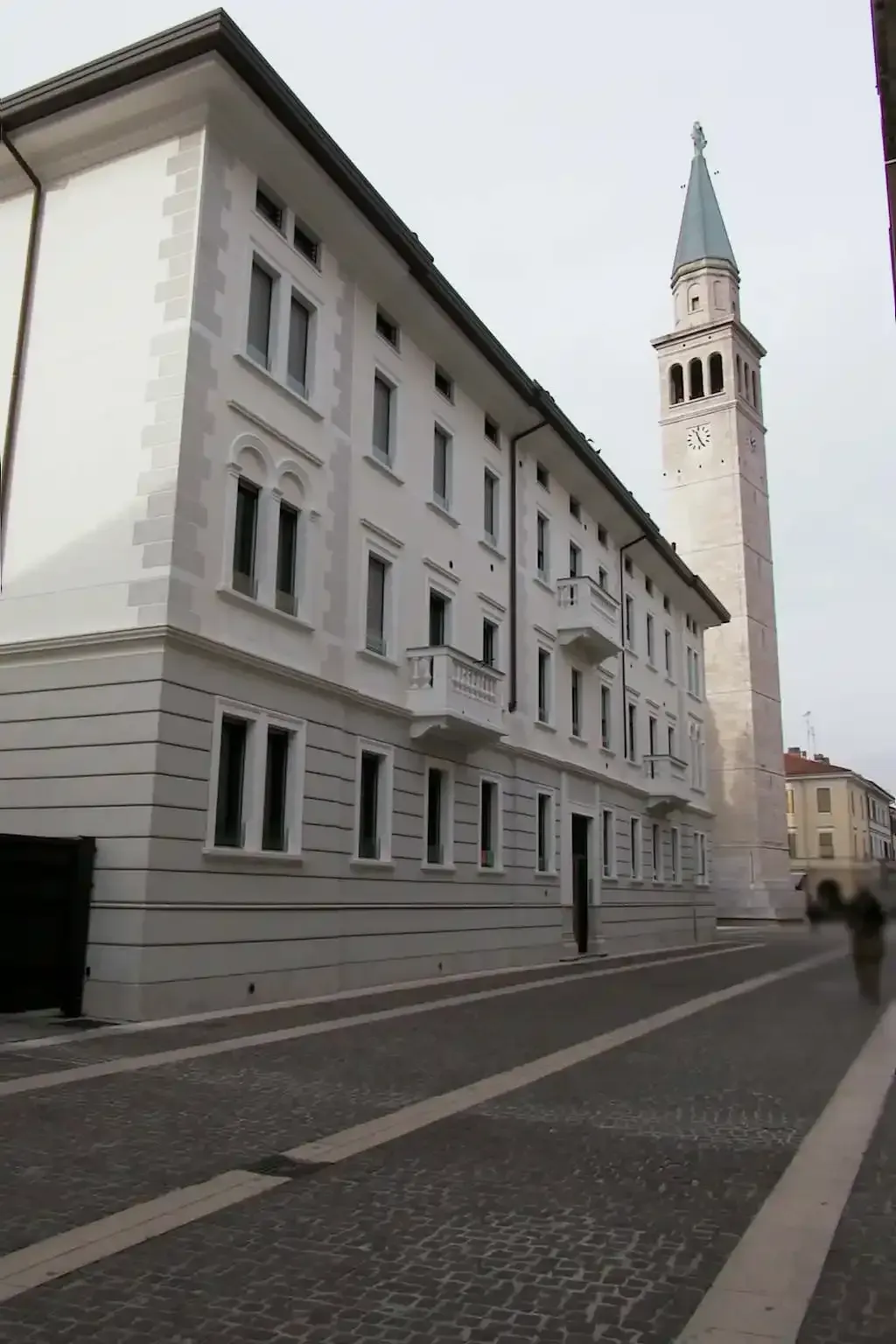 Una strada lastricata di ciottoli costeggia un alto edificio bianco a più piani e un imponente campanile in pietra nella piazza del paese.
